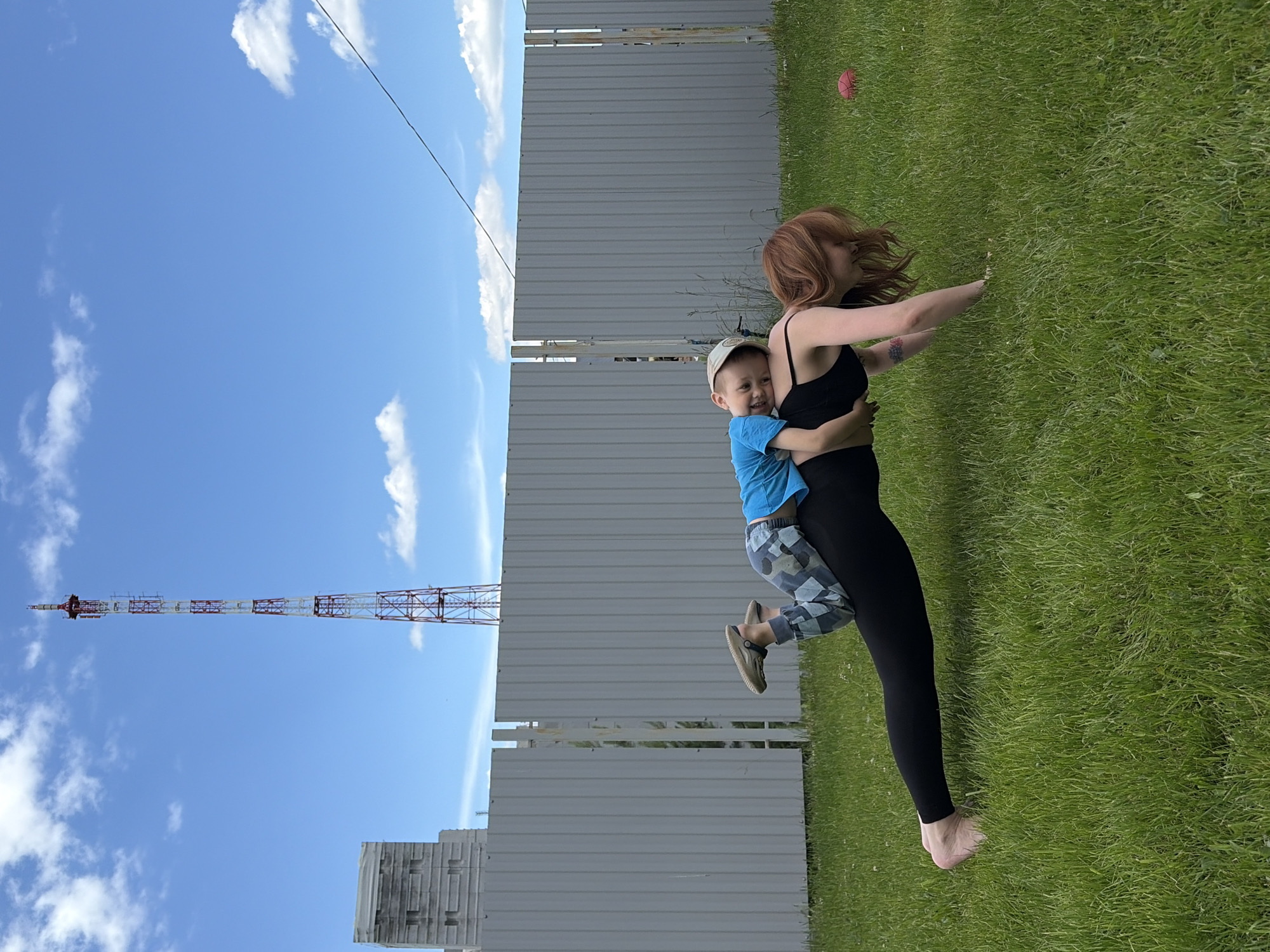 The scene captures an outdoor activity on a clear day in Chaikovsky, Perm Krai, Russia. In the foreground, a woman is positioned on a grassy lawn, performing a plank-like exercise, supporting herself on her forearms and toes. She has reddish-brown hair and is wearing a black sports bra and black leggings; a tattoo is visible on her left upper arm. A young boy is lying on her back, resting his head on her left shoulder, smiling towards the camera. He is dressed in a light blue t-shirt, camouflage-patterned pants, and light-colored slip-on shoes.
The mid-ground is dominated by a gray corrugated metal fence that runs horizontally across the frame, with a vertical gap in the center revealing some greenery behind it. In the background, under a bright blue sky with scattered white clouds, a tall, slender red and white lattice telecommunications tower is prominently visible. This tower is identifiable as the mobile communications tower located approximately at 56.764491, 54.120790, near 26 Dekabristov Street in Chaikovsky. To the left of the tower, a large, light-colored, multi-story industrial-style building, consistent with structures belonging to the Chaikovsky Textile Factory complex, is present. A dark utility line stretches diagonally across the upper right portion of the sky. A red ball is partially visible on the grass in the far right background.