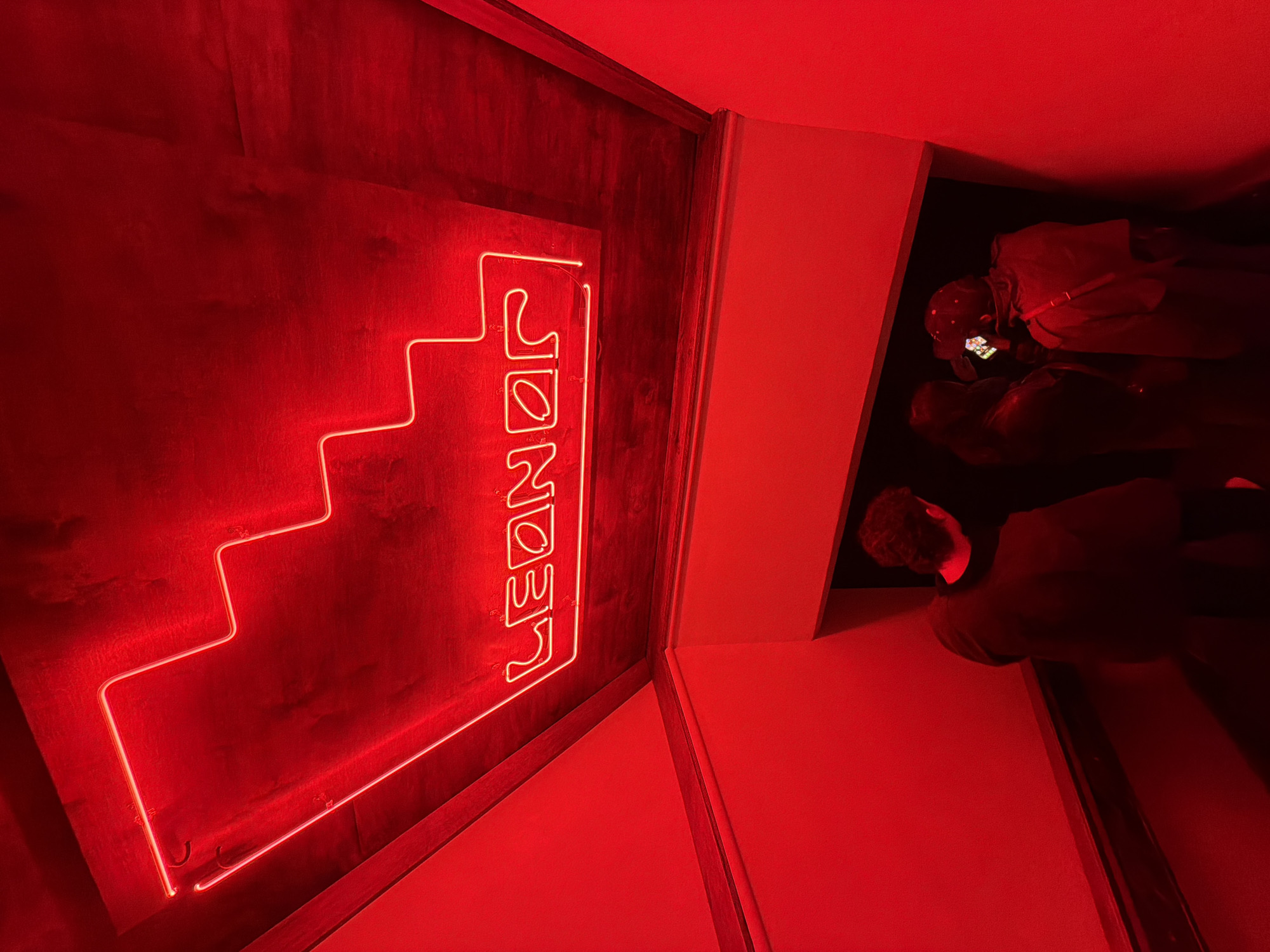 An indoor scene, saturated with red light, depicts a red neon sign on a wooden wall that reads "LEONOE" vertically, accompanied by a simple staircase design also in red neon. The sign appears to be located in a narrow hallway or doorway. In the adjacent space, partially visible, are a few people. One individual, seen from the back, is facing into the darker space. The location is in Montreal, Canada.