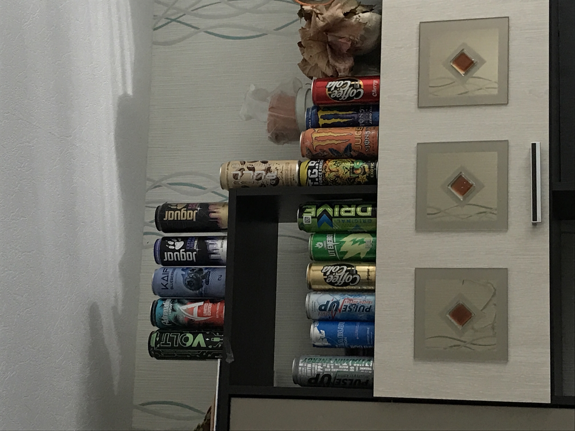 A cluttered shelf within a room contains a variety of energy drink cans stacked haphazardly. The cans display different brands and flavors, including 'Jaguar,' 'Volt,' 'Adrenaline Rush,' 'Kaif,' 'Pulse Up,' 'Lit Energy,' 'Drive,' 'Monster Juice,' and 'Coffee Cola.' Above the cans, there is a wilting floral arrangement and a package containing an indistinct item. Adjacent to the shelf, a cabinet door is visible, adorned with three square decorative panels featuring a central diamond shape with surrounding abstract lines. A thin, dark handle runs vertically along the edge of the cabinet door. The wall behind the shelf is light-colored and patterned with subtle, wavy lines. The adjacent wall to the left is a solid light gray.