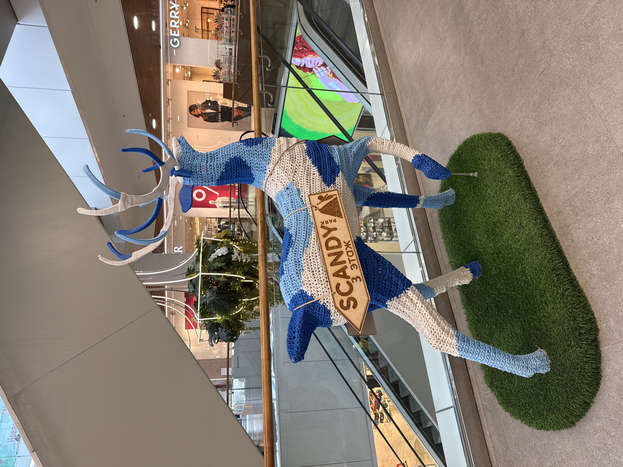 The image shows a large, crocheted deer sculpture in a shopping mall. The deer is primarily blue and white, with intricate patterns. A wooden sign is attached to its neck, reading "SCANDY 3 ЭТАЖ", which translates to "SCANDY 3rd FLOOR". The sculpture is standing on a patch of artificial grass. In the background, escalators and shop storefronts are visible, suggesting an indoor commercial environment. The lighting suggests it is daytime. The location of the shopping mall is not explicitly stated in the image, but the Cyrillic script on the sign indicates it is in a Russian-speaking region.