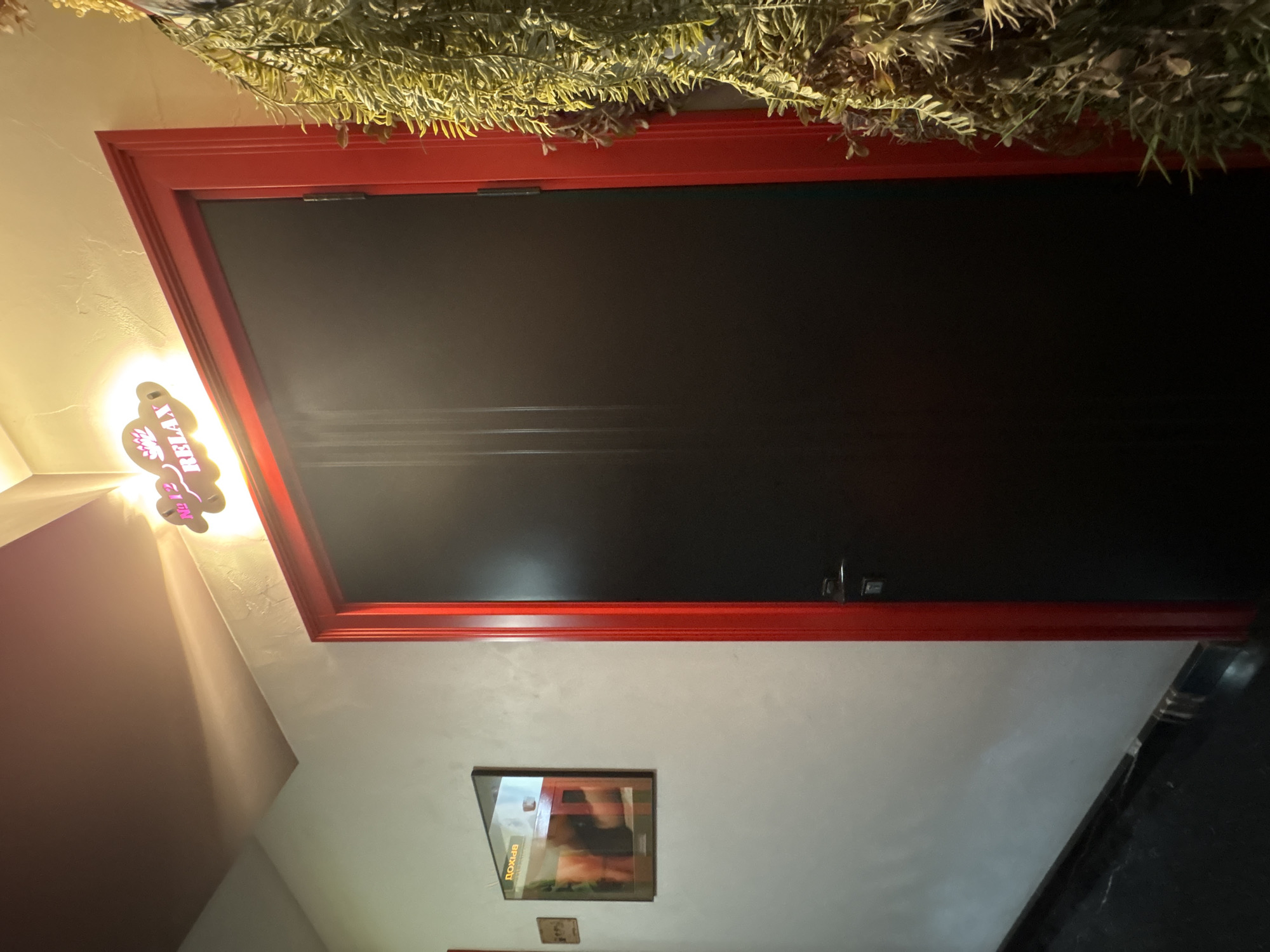 The image depicts a dark wooden door with a bright red frame, suggesting it might be an interior door. Above the door, a neon sign glows, displaying the text "№ 12 RELAX" with a lotus flower graphic. To the left of the door, a framed picture hangs on the wall, displaying an image with Cyrillic text that appears to say "ДОХОД" (income). A small, dark object is also affixed to the wall to the left of the framed picture. A large bunch of artificial green foliage hangs to the right of the door, partially obscuring it. The walls are textured, and the floor is dark. The lighting suggests an indoor setting, possibly a commercial establishment like a spa or massage parlor, given the "RELAX" sign and the foliage. The mention of Saint Petersburg, Russia, suggests this location is in that city.