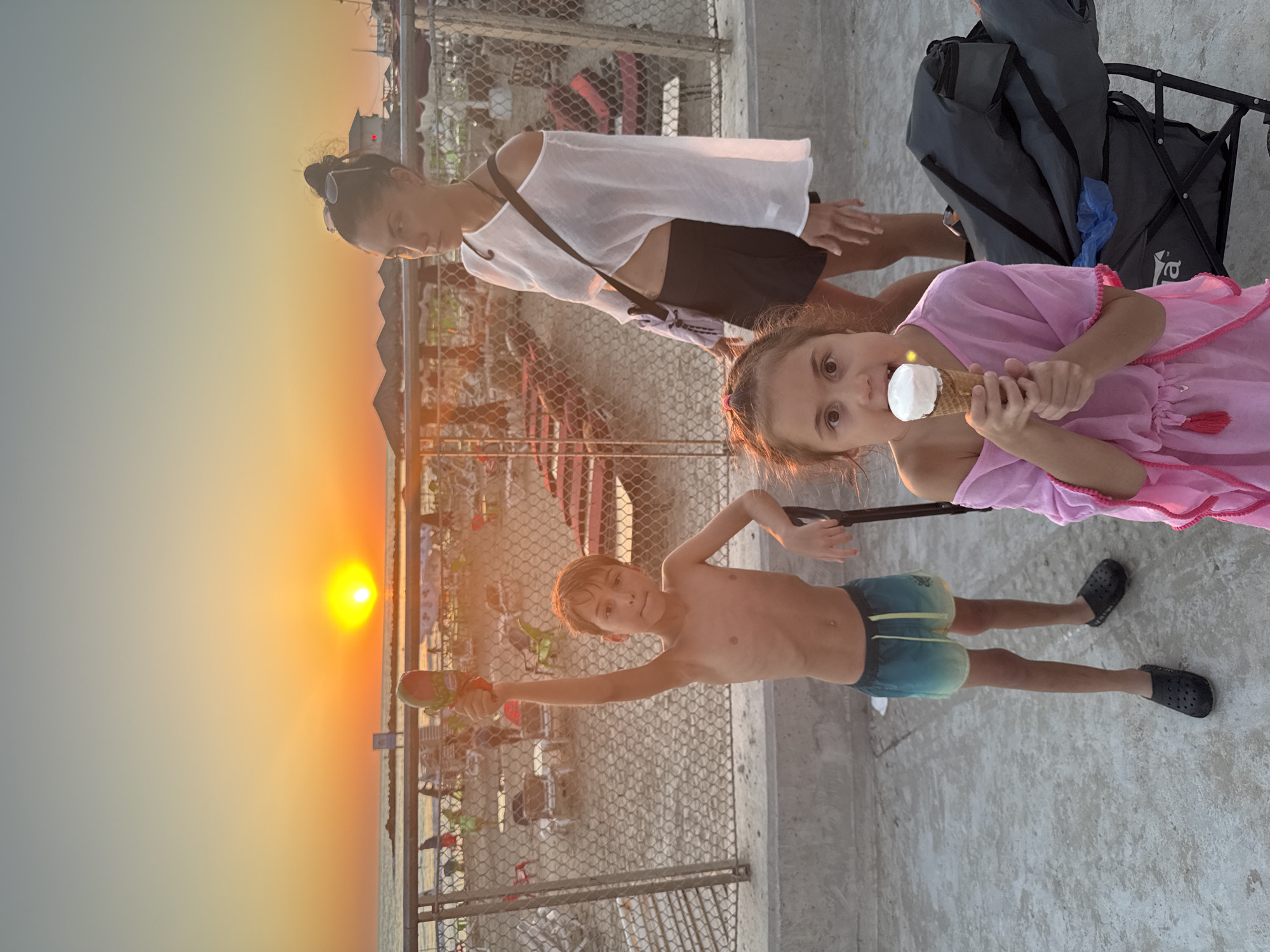 In Netanya, Israel, on a concrete surface with a view of the Mediterranean Sea, a family enjoys the sunset. A boy stands on the left, shirtless and wearing ombre swim trunks, holding a popsicle aloft in his right hand. To the right, a woman wears a white, off-the-shoulder top and a black skirt, with a bag slung across her body. In the foreground, a young girl in a pink cover-up eats an ice cream cone. A dark gray wagon is parked behind the girl. The background features a beach with numerous lounge chairs and umbrellas, indicating a public beach area. The sun is low on the horizon, casting a warm orange glow over the scene.