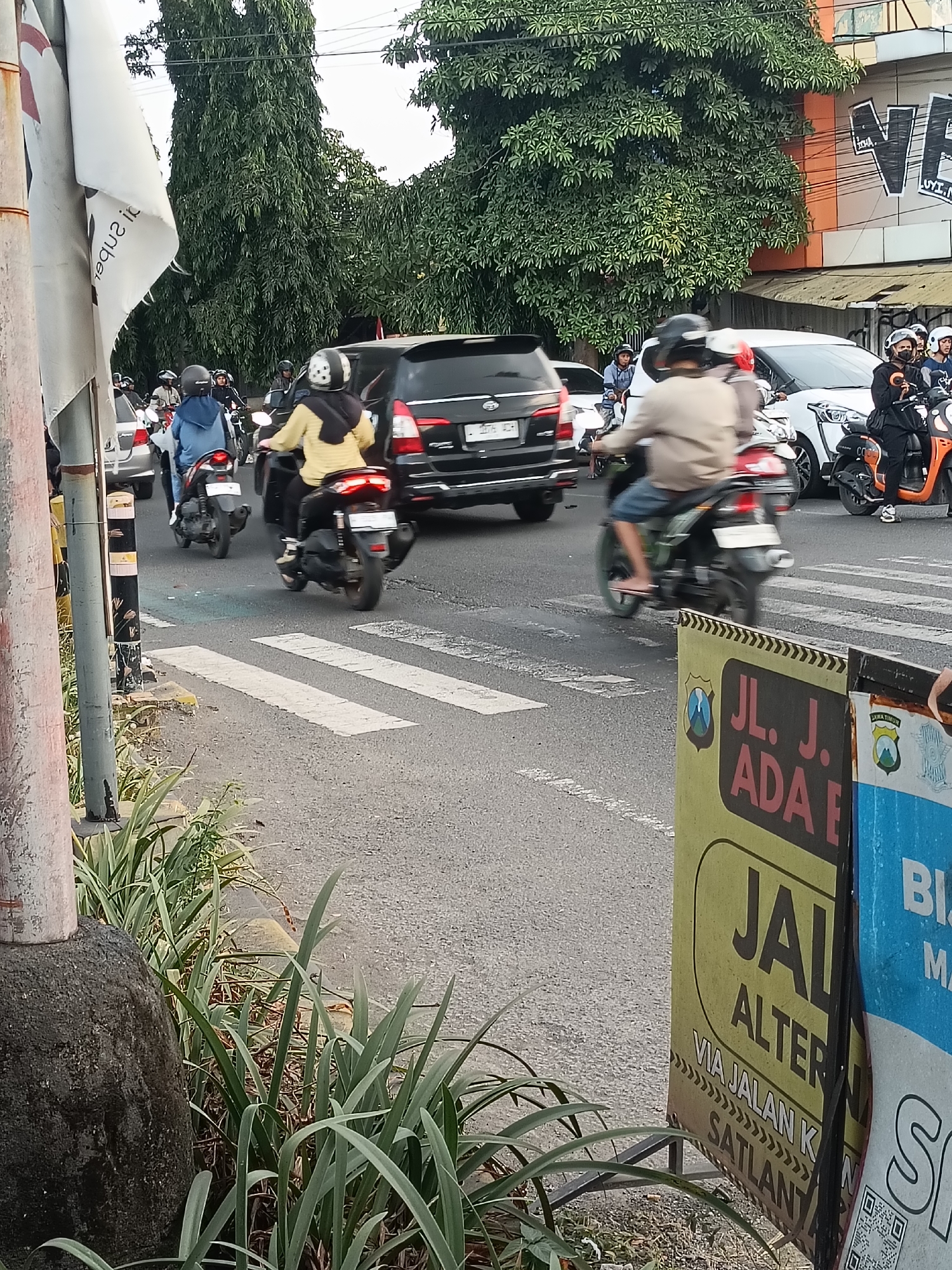 The image depicts a street scene in the city of Kediri, Indonesia, featuring a busy intersection with multiple motorcycles and cars. A crosswalk is visible on the asphalt road. Several individuals are riding motorcycles, some wearing helmets and backpacks. One person in the foreground is wearing a yellow shirt and a black hijab, while another wears a blue jacket and a blue headscarf. A black SUV is prominent in the middle of the frame, and a white car is positioned to its right. Traffic appears to be moderate. In the background, lush green trees line the street, and buildings are visible. A vertical pole with black and white stripes stands on the left side of the image. On the right, several signs are posted, including one with text in Indonesian that reads "JL. J. ADA E" and another with "JAL ALTER" and "VIA JALAN K SATLAN." These signs suggest this is likely a public road within Kediri. The overall atmosphere is that of a typical urban traffic environment in Indonesia.