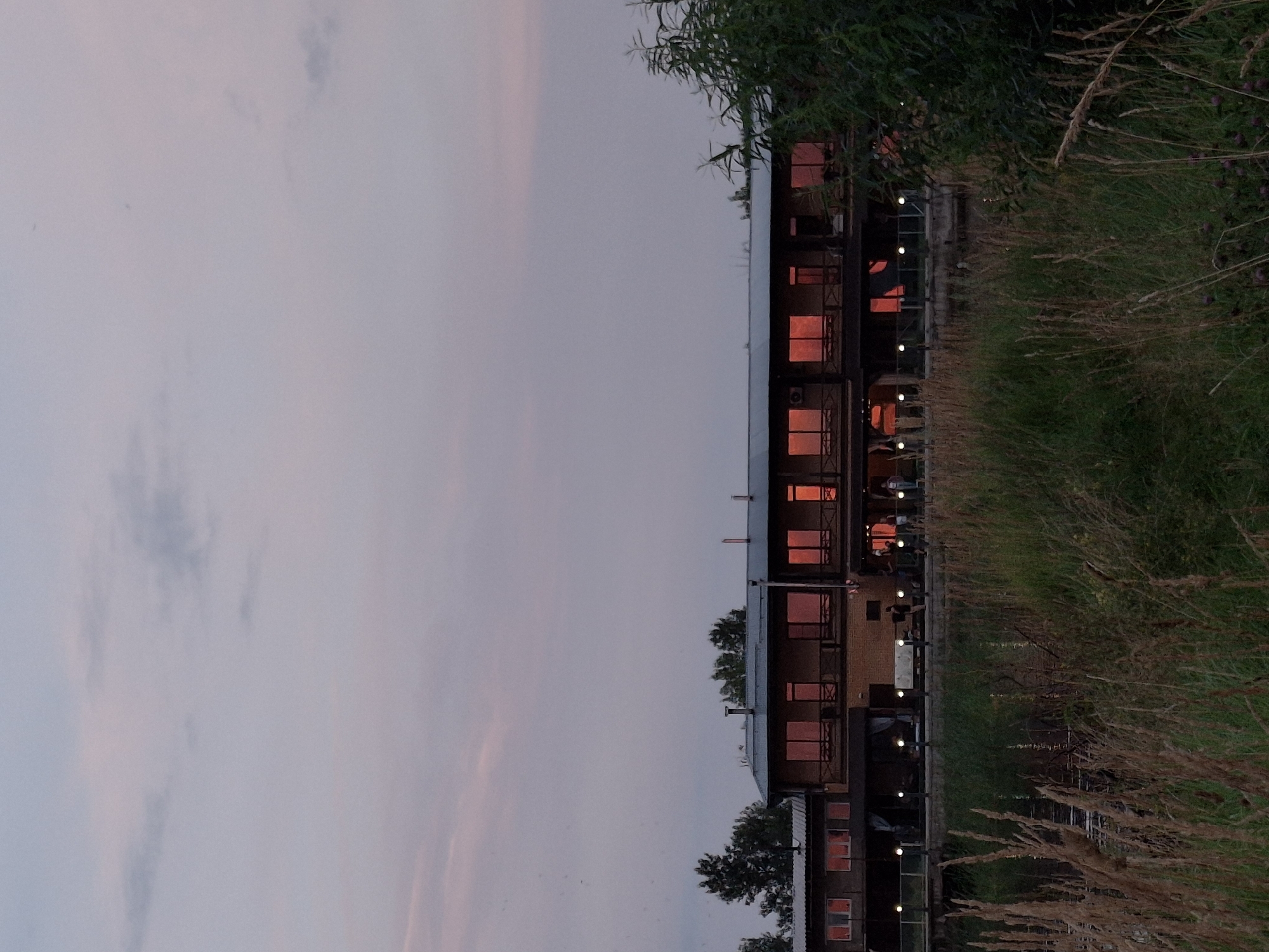 The image depicts a twilight scene in Chaikovsky, Russia, featuring a two-story building with a light brown brick facade, possibly a restaurant or hotel, situated on the banks of the Kama River. The building has numerous large windows that reflect the warm hues of the sunset, casting orange and pink glows. Several people are visible on the building's terrace, appearing to be casually strolling or conversing. The foreground is dominated by tall, dry grass and reeds, suggesting a natural, undeveloped area along the riverbank. Trees are scattered behind the building, and the sky is a soft blend of pink and gray clouds. The overall atmosphere is serene, with the natural landscape juxtaposed against the human activity at the building.