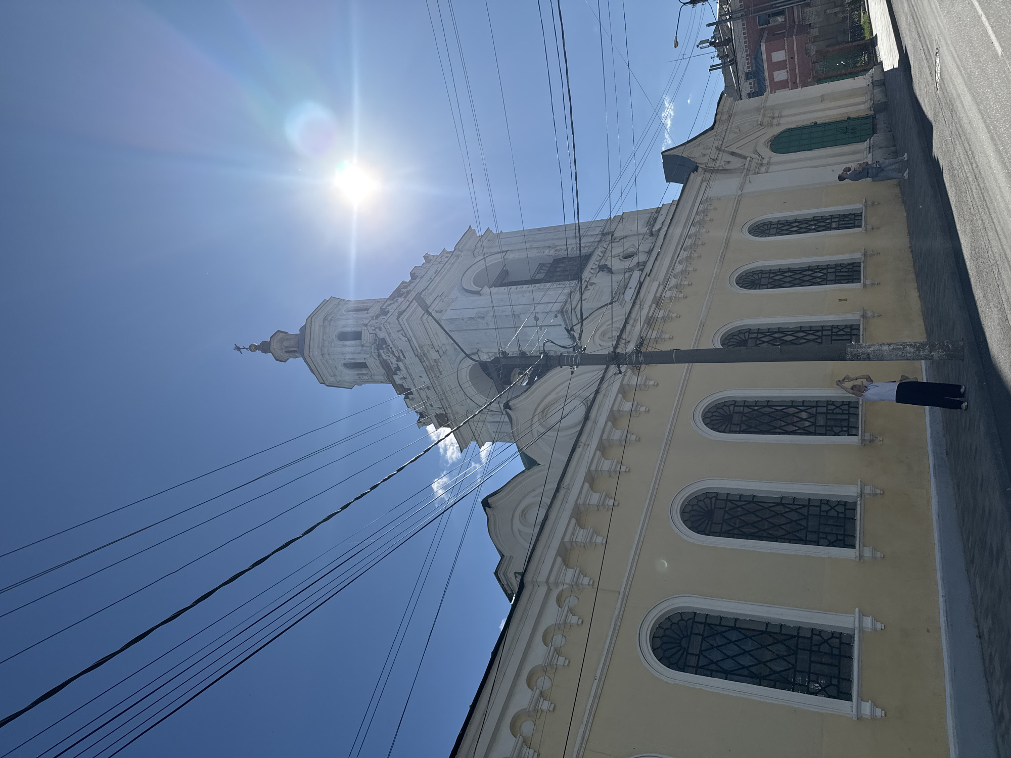 In the city of Yelets, Russia, a low-angle shot captures a pale yellow building with arched windows and white decorative elements. To the right, a white bell tower with arched openings and a cross at its apex rises against a bright blue sky. The sun is prominently visible, creating a lens flare. Overhead, a network of dark electrical wires crisscrosses the frame. On the sidewalk in front of the building, a person in dark pants and a light shirt stands facing away from the camera, with one arm raised. Further to the right, two people, one holding a child, are visible near a bright green door. The scene includes a paved street in the foreground.