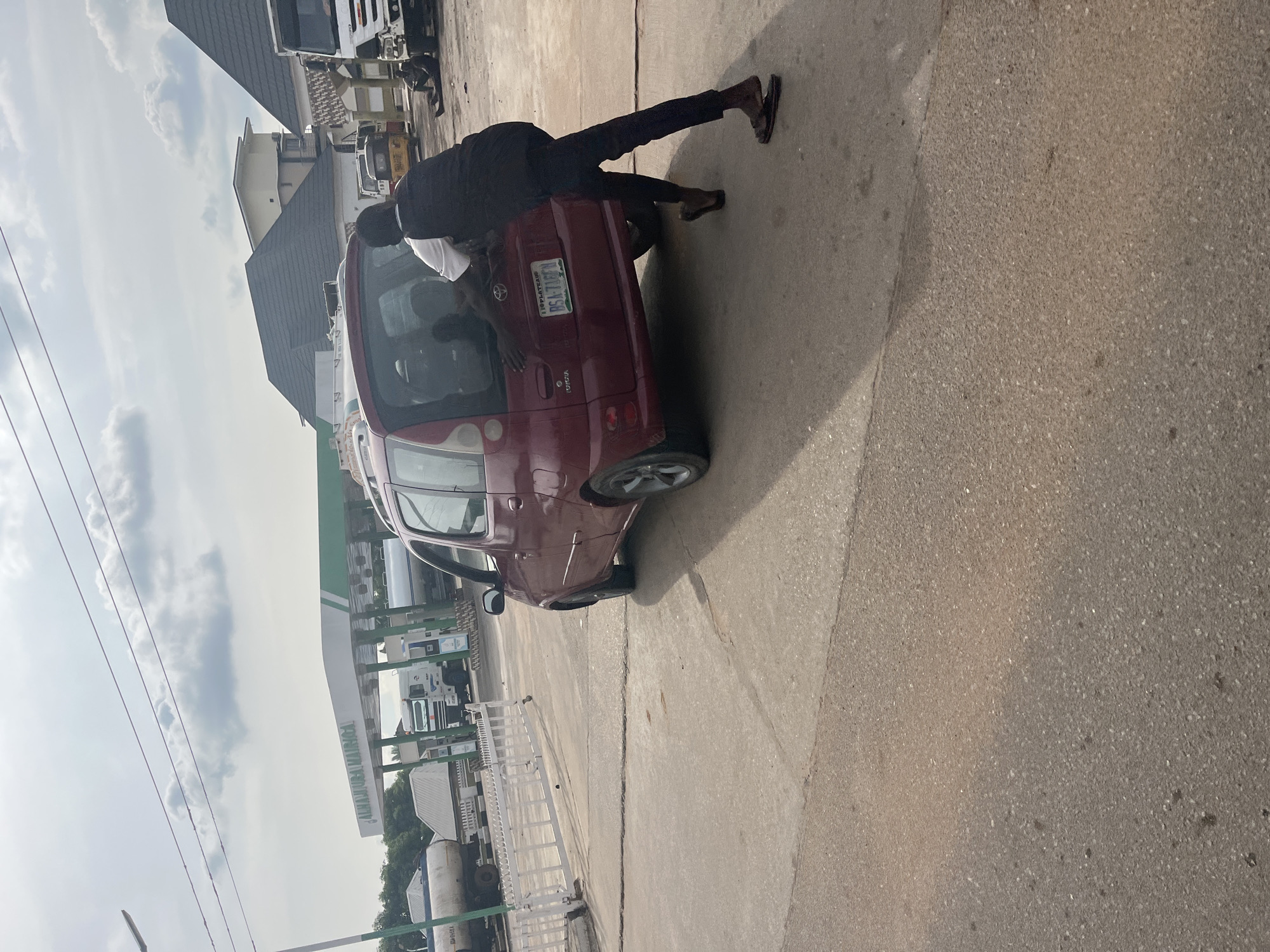 A man is pushing a red minivan at a gas station in Jalingo, Nigeria. The minivan is positioned facing away from the camera, and the man is on the right side of the vehicle, pushing it from behind. The man is wearing a white t-shirt and dark pants, and his feet are bare except for flip-flops. The gas station has a canopy with the words "ALHAJI UBA MAIRIGA" visible. In the background, there are several fuel tankers, a white truck, and some buildings with tiled roofs. The sky is partly cloudy. The ground is paved with asphalt and concrete.