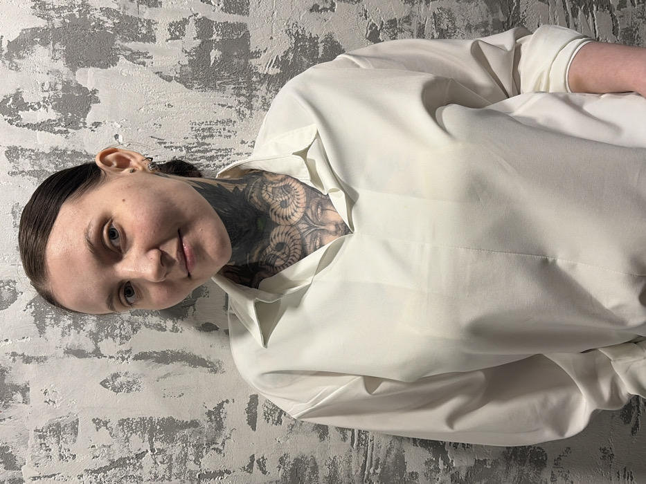 A female subject is depicted from the mid-torso up, standing against a textured wall in Chaikovsky, Russia. She has dark hair pulled back, a clear complexion, and a slight smile while looking directly at the camera. Her neck and upper chest display extensive black and grey tattoos, featuring intricate designs including coiled elements and a stylized face or figure. She wears a light-colored, possibly white or off-white, collared blouse with a V-neck, and long sleeves that appear to be rolled up at the cuffs. A small hoop earring and a stud are visible in her right ear. The background wall exhibits a distressed, uneven texture with varying shades of grey and white, suggesting peeling paint or rough plaster.