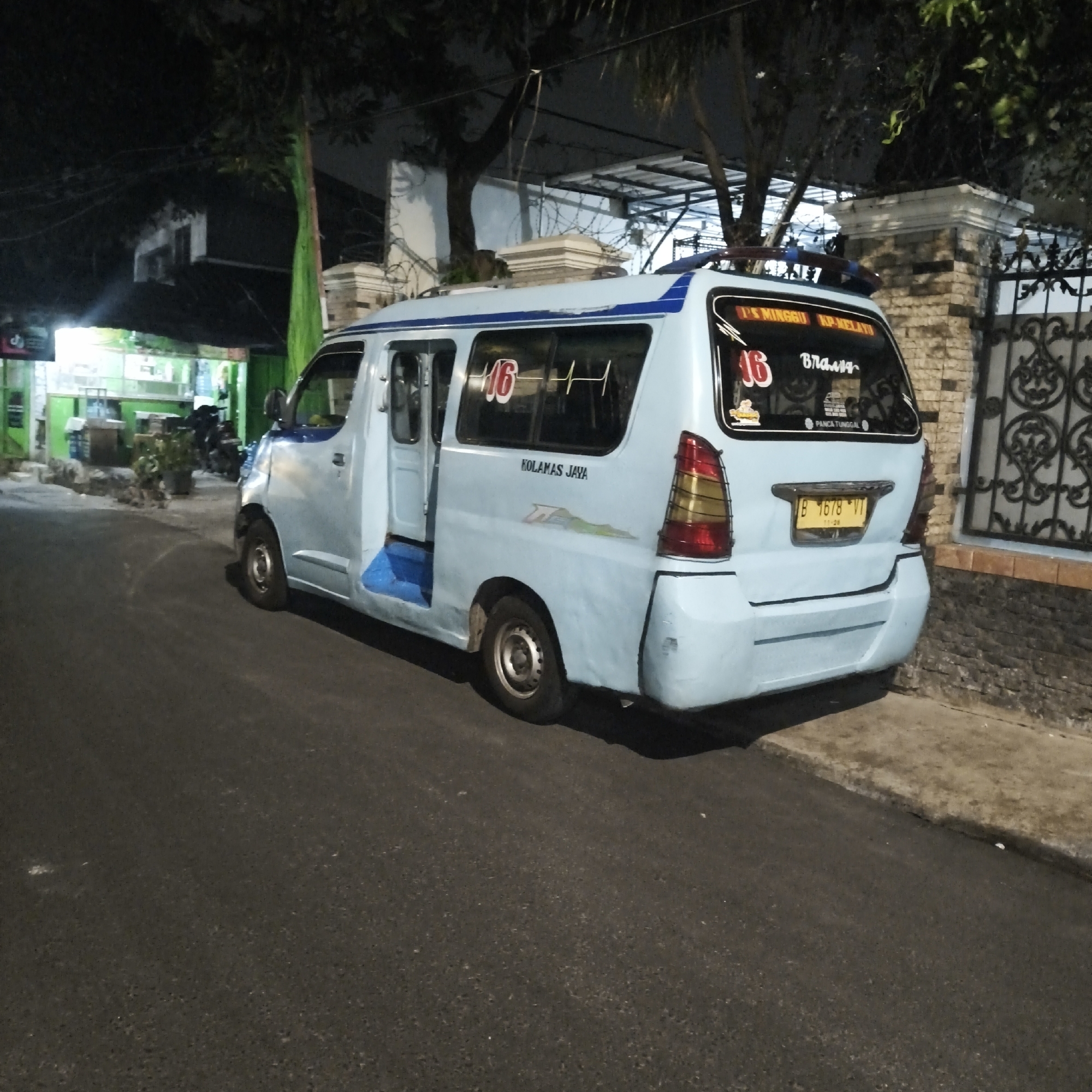 A light blue passenger van is parked on the side of a paved road at night. The van has several decals and markings on its exterior. On the rear passenger side door, the text "KOLAMAS JAYA" is visible. Above the rear window, text reads "P'S MINGGU KP-MELAYU" and "16". Another "16" is also visible on the side window. A yellow license plate with the characters "B 1678 VT" is mounted on the rear of the van. The van's doors are open, revealing blue steps inside. A brick wall with a decorative metal fence is visible behind the van, along with trees and foliage. In the background, a building with green accents and glowing lights suggests a commercial establishment. The scene appears to be in a residential or commercial area of South Jakarta, Indonesia.