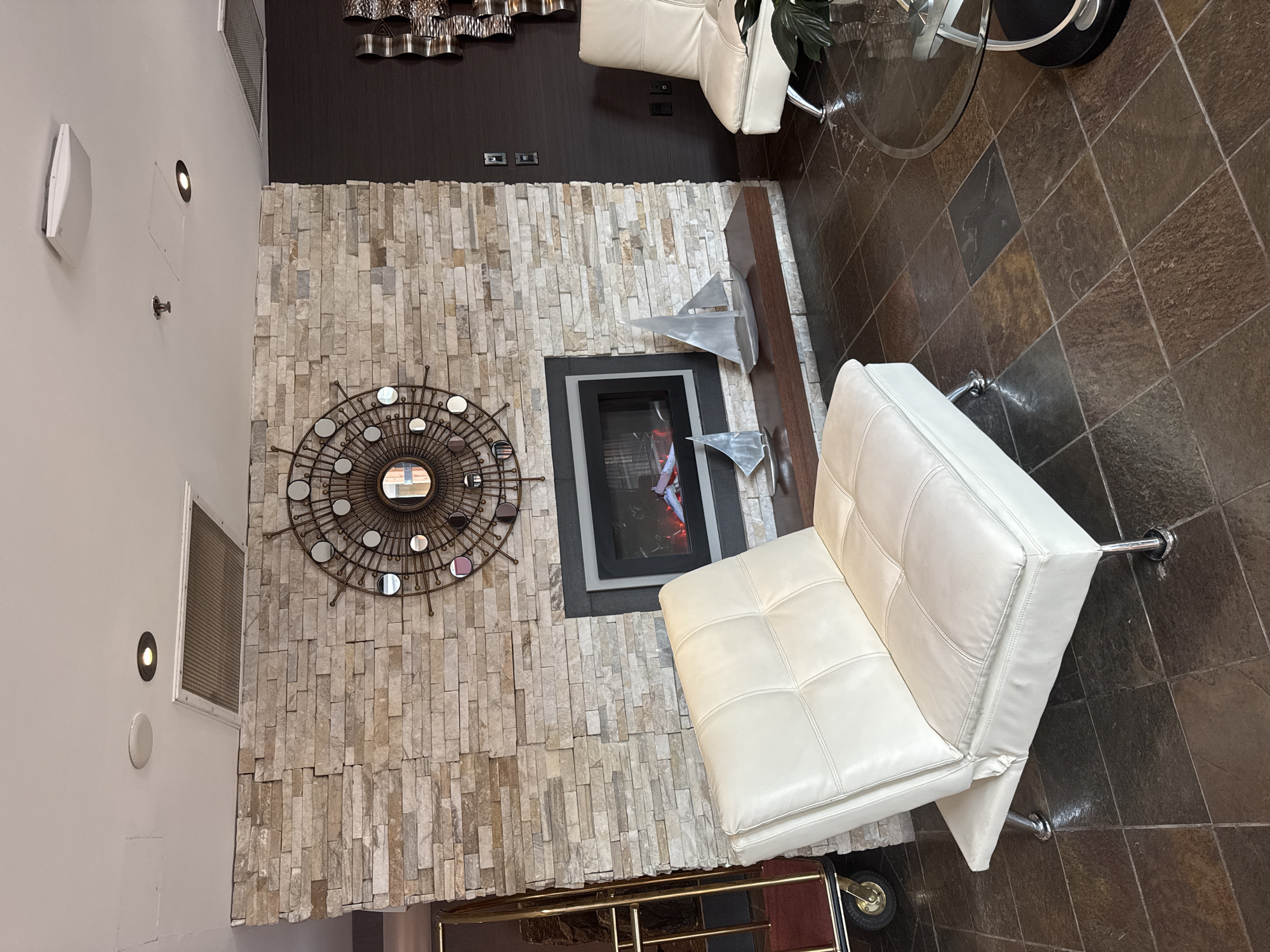 The image depicts an indoor waiting area, possibly a hotel lobby in North Vancouver, Canada. In the foreground, a white leather loveseat with silver legs is positioned on a dark, tiled floor. Behind the loveseat, a fireplace with a stone veneer facade is the central element. The fireplace is lit, showing orange flames. Above the fireplace, a large, circular decorative mirror with reflective elements is mounted. To the right of the fireplace, on a dark wooden shelf, are two silver sailboat figurines. Further to the right, a white armchair with silver legs is visible, along with a glimpse of a glass-top coffee table. To the far left of the image, a golden luggage cart with a red mat is partially visible. The walls are a mix of stacked stone and dark textured wallpaper. The ceiling features recessed lighting and ventilation grates.