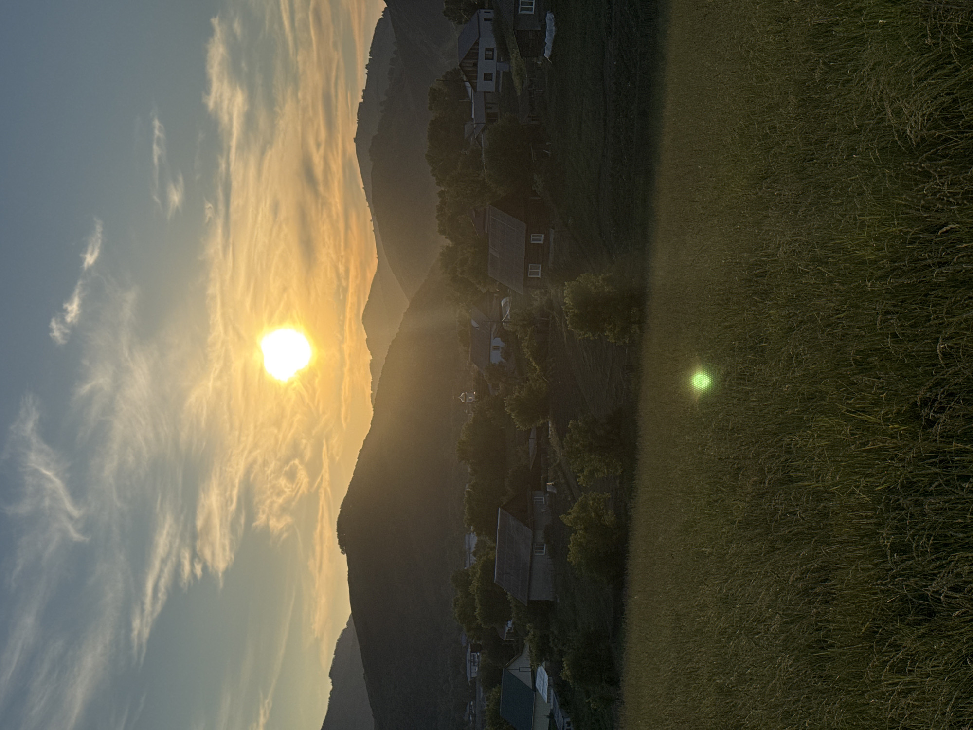 The image depicts a rural village scene in Soloneshnoye, Russia, at sunset. Rolling hills covered in trees form a dark silhouette against the bright, sunlit sky. The sun is a brilliant, round orb positioned in the upper center of the frame, casting a warm, golden glow across the landscape. Wispy clouds are scattered across the blue sky. In the midground, nestled among trees, are several houses with pitched roofs, indicative of a village. Some houses appear to be wooden, while others have lighter colored walls. A patch of tall, green grass dominates the foreground, with a subtle, bright green lens flare visible in the lower center of the image. The overall atmosphere is serene and tranquil, characteristic of a rural sunset.