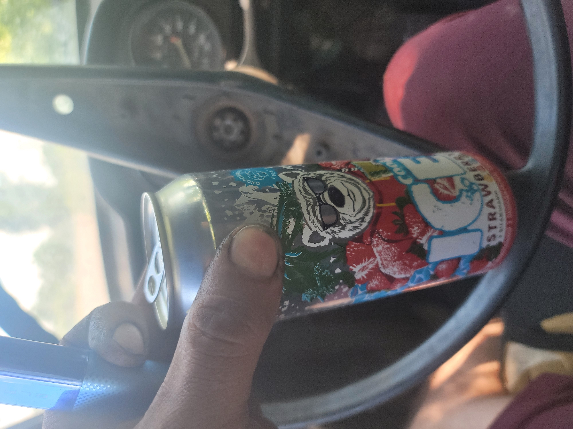 The image shows a close-up of a person's hand holding a can of "Cool Ice Strawberry" drink inside a vehicle. The person is wearing dark-colored fingernails and is holding the can with their right hand. The can has a drawing of a bear wearing sunglasses on it, along with images of strawberries. The background is blurred, but it appears to be the interior of a car with a steering wheel and dashboard visible. The overall lighting suggests it is daytime. The image does not provide any specific details that would indicate its location within Pereshchepyne, Ukraine.