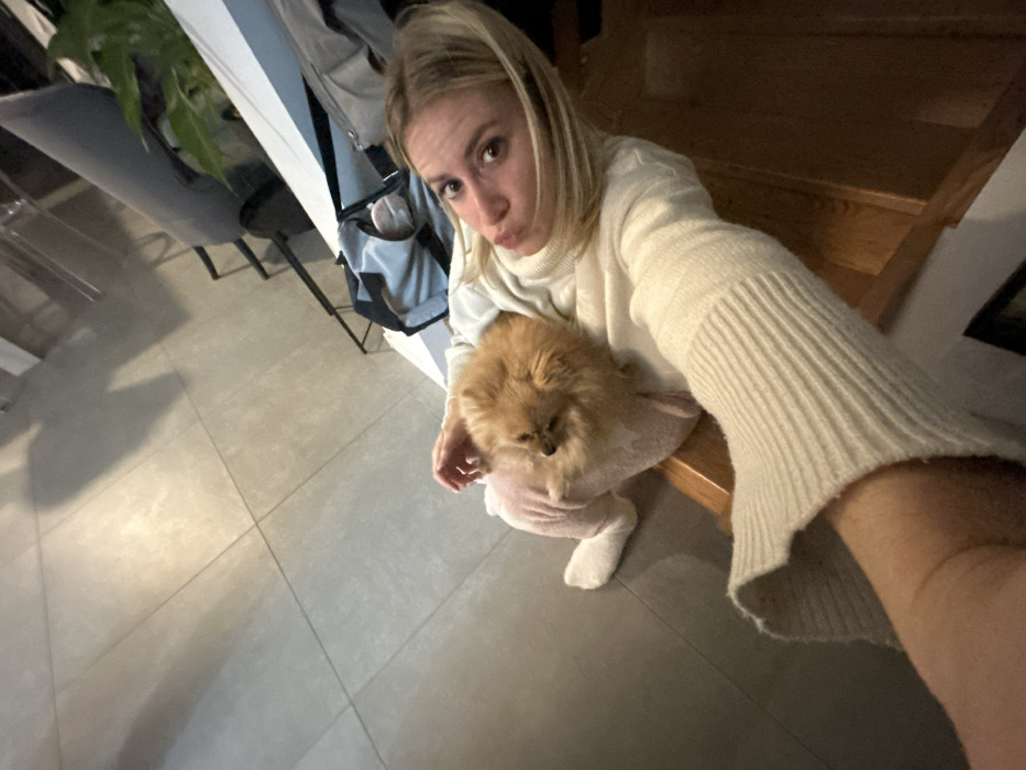 A blonde woman in a cream sweater takes a selfie. She holds a small, fluffy, brown dog wearing a pink sweater. The background includes a grey tiled floor, a grey chair, a white wall with coats hanging on it, and a staircase. The lighting is somewhat dim, giving the photo a cozy indoor feel. The woman is pouting, adding a playful element to the scene.