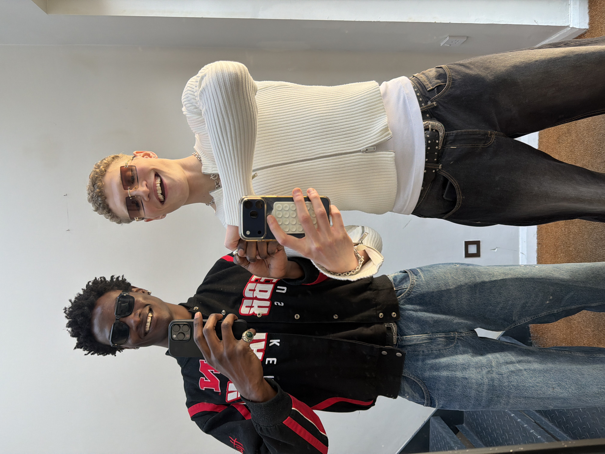 Two young men are posing for a mirror selfie. The man at the top of the frame is light-skinned with short blond hair and is wearing sunglasses, a white zip-up sweater, a white t-shirt, a silver necklace, and dark jeans with a studded belt. He is holding a gold smartphone with his right hand. He is smiling. The man at the bottom of the frame is dark-skinned with black hair and is wearing sunglasses and a black bomber jacket with red stripes and white lettering, and blue jeans. He is holding a black smartphone with his right hand. He is smiling. The location appears to be indoors, possibly a store or studio, with a white wall in the background. The exact location in the city of London, United Kingdom cannot be determined from the visual information.