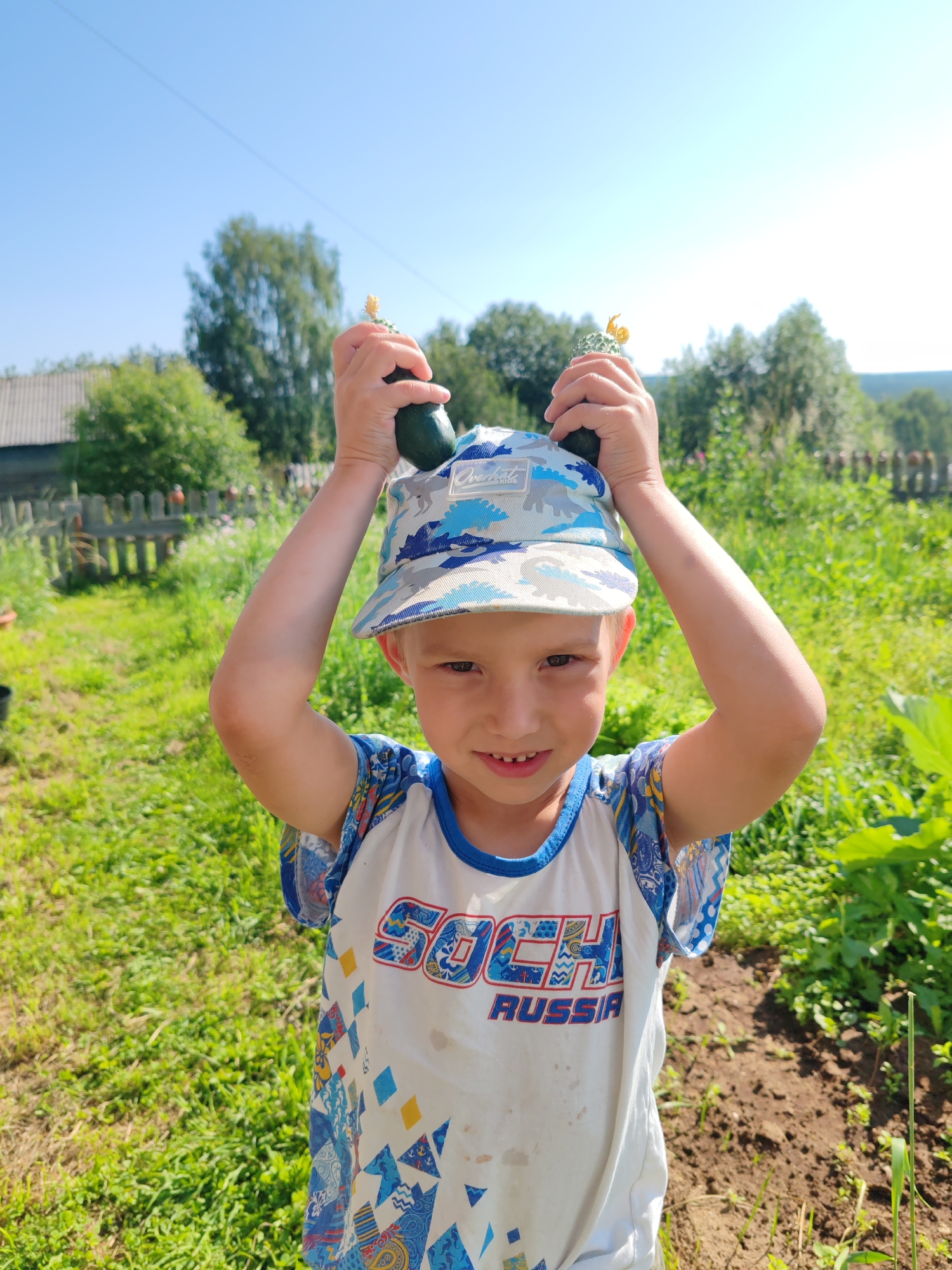 A young Caucasian boy with blond hair and fair skin is pictured outdoors, holding two small cucumbers above his head. He is wearing a white t-shirt with blue trim and a camouflage baseball cap. The t-shirt has the word "SOCHI RUSSIA" printed on it. The boy is smiling and looking at the camera. The background shows a sunny, rural landscape with green grass, trees, and a wooden fence. The scene appears to be in the village of Chuchlema, Russia. The boy's posture and the cucumbers suggest he is either showing off a harvest or playing a game.
