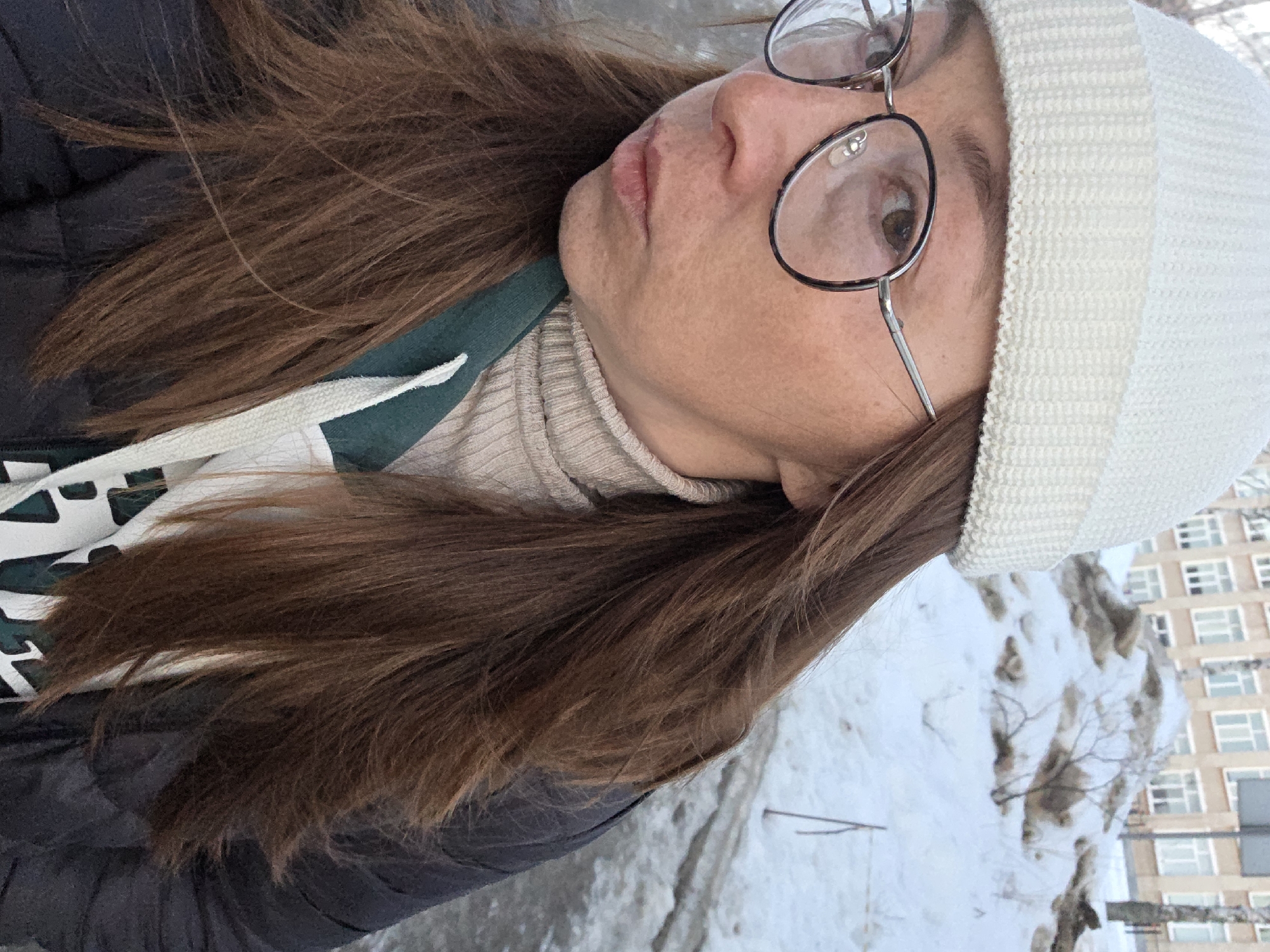 The image features a female subject in the city of Chaikovsky, Russia, in an outdoor winter or early spring setting. The subject, viewed from the chest up and slightly angled left, is looking towards the camera. She wears a white knitted beanie hat, round metal-framed glasses, a beige ribbed turtleneck sweater, and a dark, possibly black, quilted jacket. Her long, straight, medium-brown hair is visible over her shoulders and chest. A dark green strap from a bag, with a white accent, crosses her left shoulder. Her right eye, with a brown iris, is clearly visible, while her lips are closed and slightly pursed. The background consists of a snow-covered environment. A substantial, dirty snow pile with bare branches protruding from it is visible behind the subject. Further in the background, a light-colored, multi-story building with numerous rectangular windows can be observed. The ground immediately behind the subject is a mix of dirty snow and exposed pavement. A thin, dark stick stands vertically in the snow on the far right of the frame.