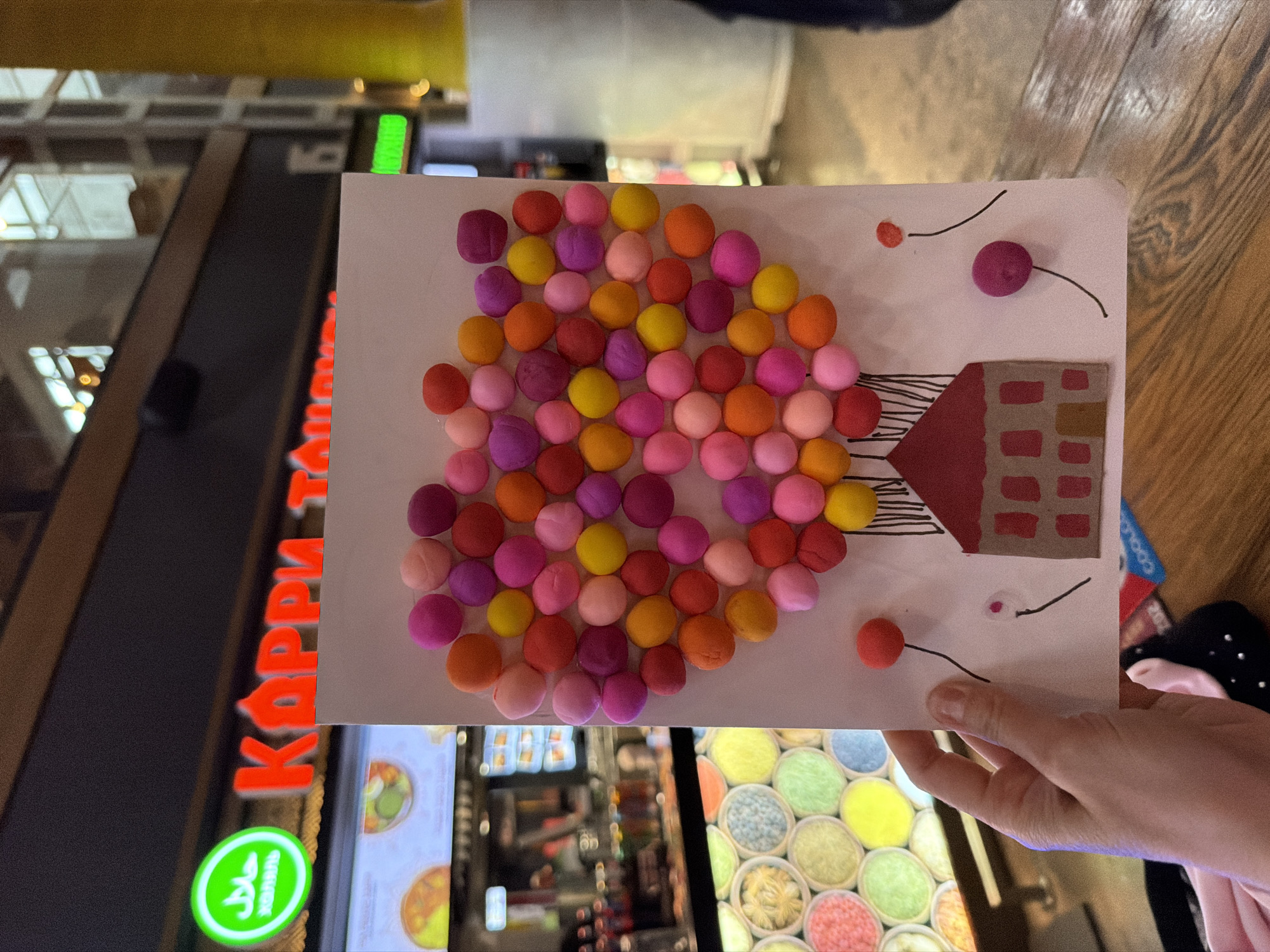A child's artwork is being held up in front of a retail establishment. The artwork depicts a simplified house suspended by balloons of varying colors, constructed from modeling clay adhered to a white paper. The background reveals the storefront of a business named "КАРРИ ТАЙ" with a green "Halal" logo, indicative of a restaurant selling halal Thai curry. This business is located in Moscow, Russia, due to the Cyrillic lettering on the store sign. The overall environment suggests an indoor retail space within the city. A person is holding the artwork. The background blur reveals a display of small containers with colored contents (potentially candies or food toppings), along with general interior elements of the storefront.