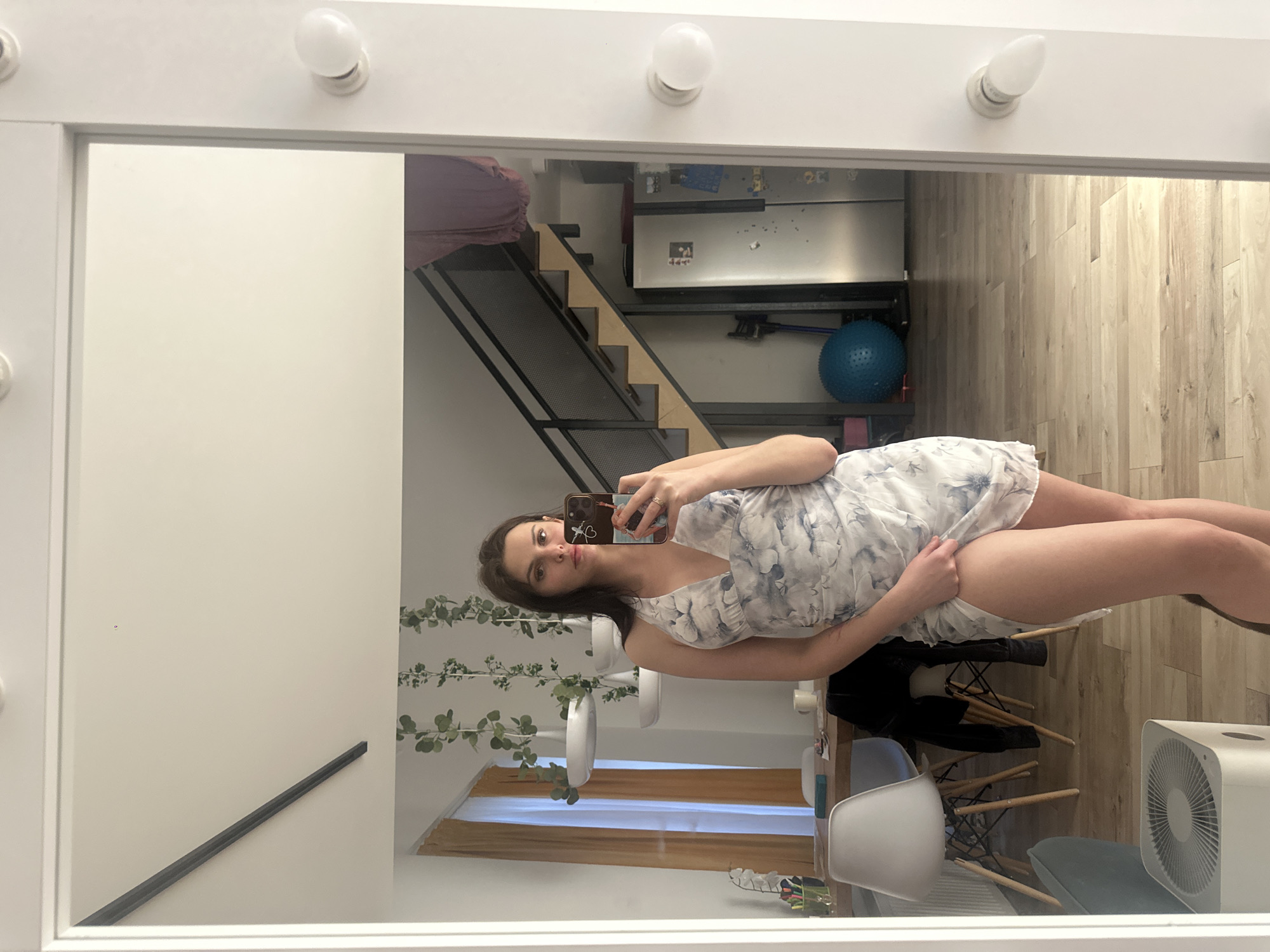 A young woman with dark hair is taking a selfie in a mirror. She is wearing a short, halter-neck dress with a blue and white floral pattern. She is standing in a room that appears to be a living area or dining room. In the reflection, a dining table with chairs, a staircase, and a refrigerator are visible. The lighting suggests it is daytime.