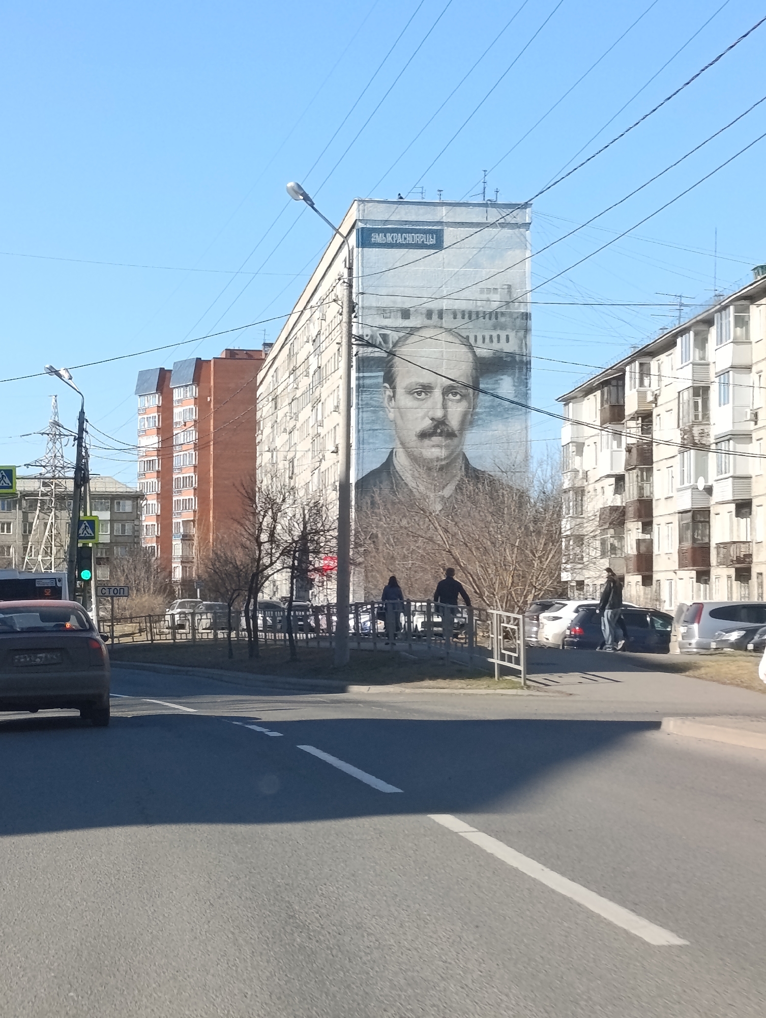 The image shows a street scene in Krasnoyarsk, Russia. In the background, a large mural of a man with a mustache is painted on the side of a tall, gray apartment building. Above the mural, text in Russian reads "#МЫКРАСНОЯРЦЫ" (We are Krasnoyartsians). Other apartment buildings of varying heights and colors are visible in the distance. A traffic light with a green light is visible on a pole. Several cars are parked along the street, and pedestrians are present on the sidewalk near a metal railing. The foreground shows a gray car driving on a paved road with white lane markings. The sky is blue.