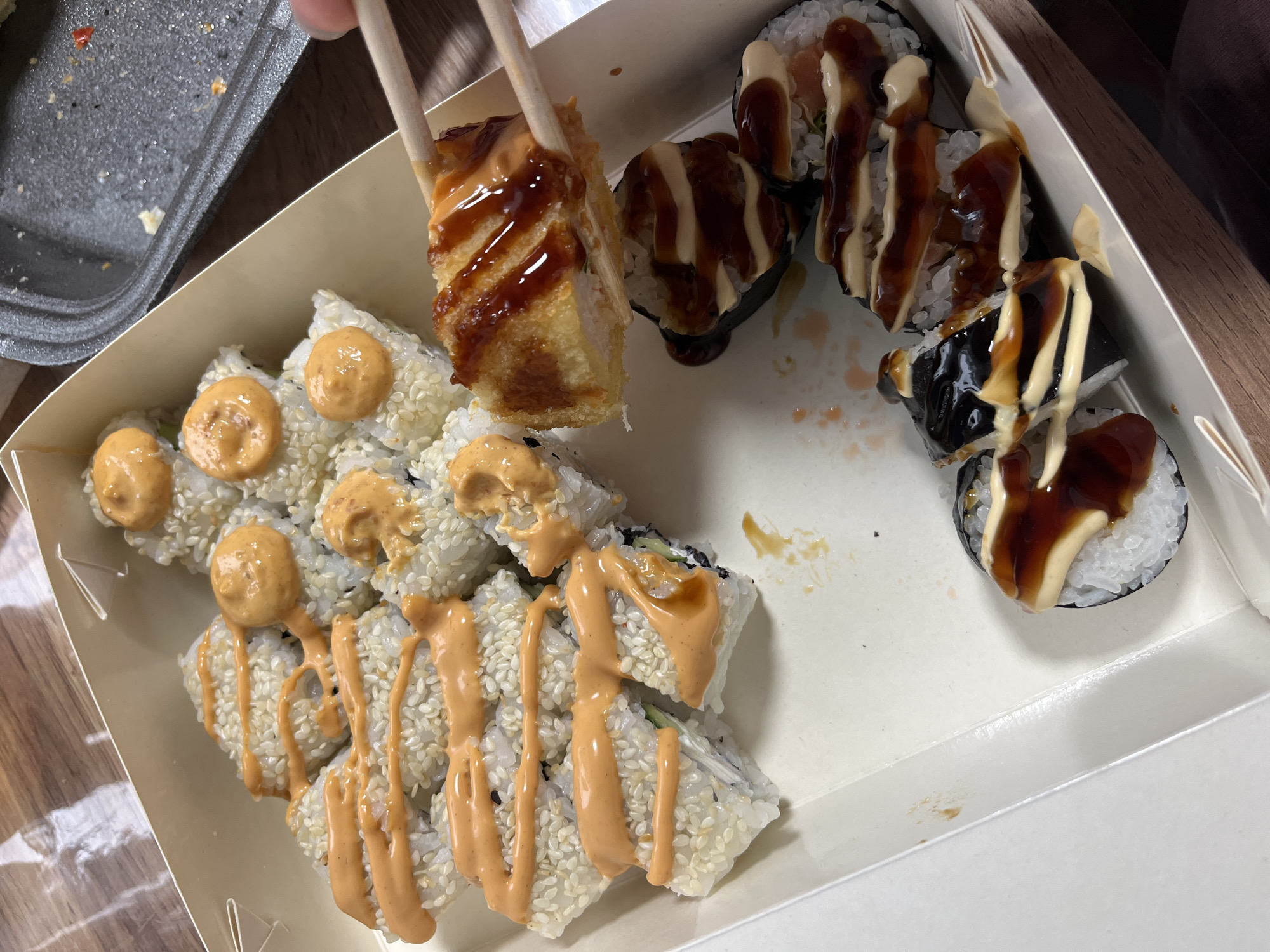 The image shows a close-up of a white cardboard box filled with sushi. In the foreground, there are several pieces of sushi rolls covered in white sesame seeds and drizzled with a creamy orange sauce. Another set of sushi rolls in the background are darker, wrapped in seaweed and topped with a combination of dark brown and white sauces. One piece of sushi has been picked up with chopsticks and is being held above the box, revealing its fried exterior and a drizzle of dark sauce. The sushi appears to be part of a meal, likely for takeout or delivery, given the disposable container.
The setting is indoors, with a wooden surface visible in the background. There are no people clearly visible, though hands holding chopsticks suggest someone is about to eat the sushi. The lighting suggests it could be daytime, but there are no strong indicators of time or weather. The presence of sushi suggests a casual dining experience or a meal at home. No text is visible in the image. The location context of Nizhny Tagil, Russia, provides a geographical setting for this image.