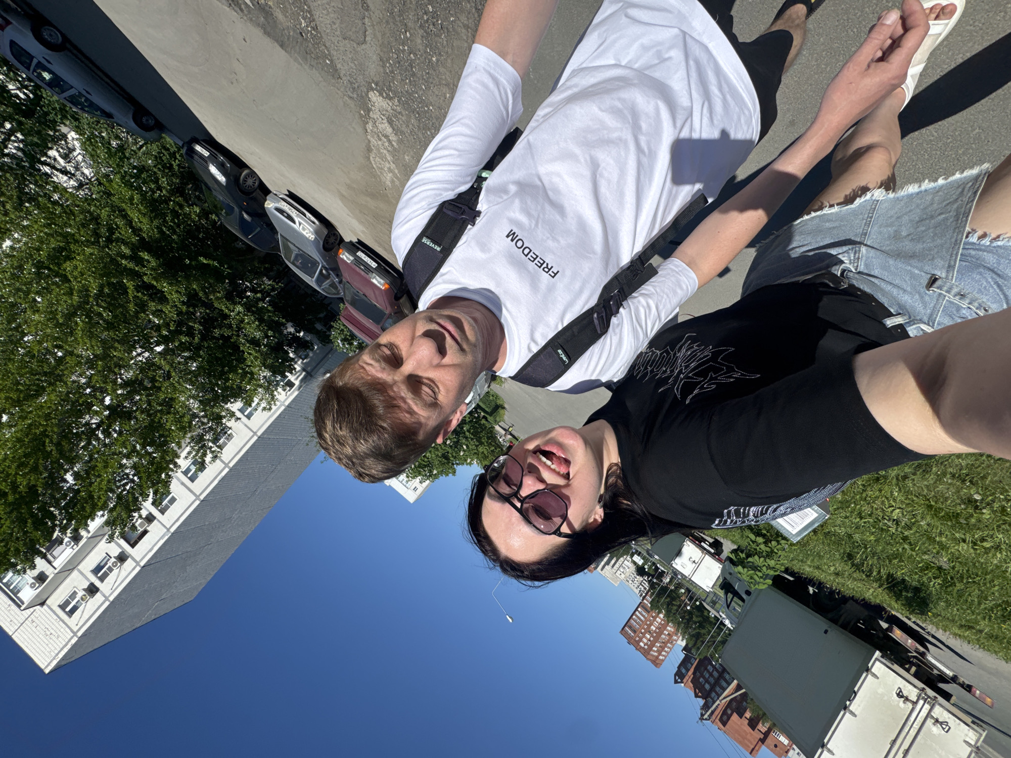 A selfie captures a fair-skinned man and a woman with long black hair and sunglasses, taken outdoors in daylight, presumably in the city of Unknown, Unknown. The man is wearing a white long-sleeved shirt with "FREEDOM" printed on it, while the woman is in a black shirt and denim skirt. Their hands are lightly clasped. The background features a blend of urban and natural elements, including multi-story buildings, a clear blue sky, trees, parked cars, and delivery trucks, suggesting an urban setting with nearby residential areas.