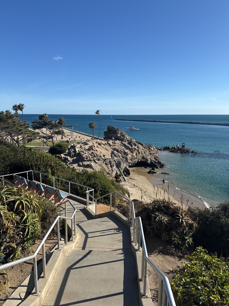 The image shows a view of the ocean and beach from a set of concrete stairs with metal railings. The location is Newport Beach, United States. The sky is clear and blue. The beach is sandy, and there are some people walking on it and swimming in the water. There are palm trees and other greenery along the coast. A rocky outcrop is in the middle ground. A breakwater extends into the ocean in the background.