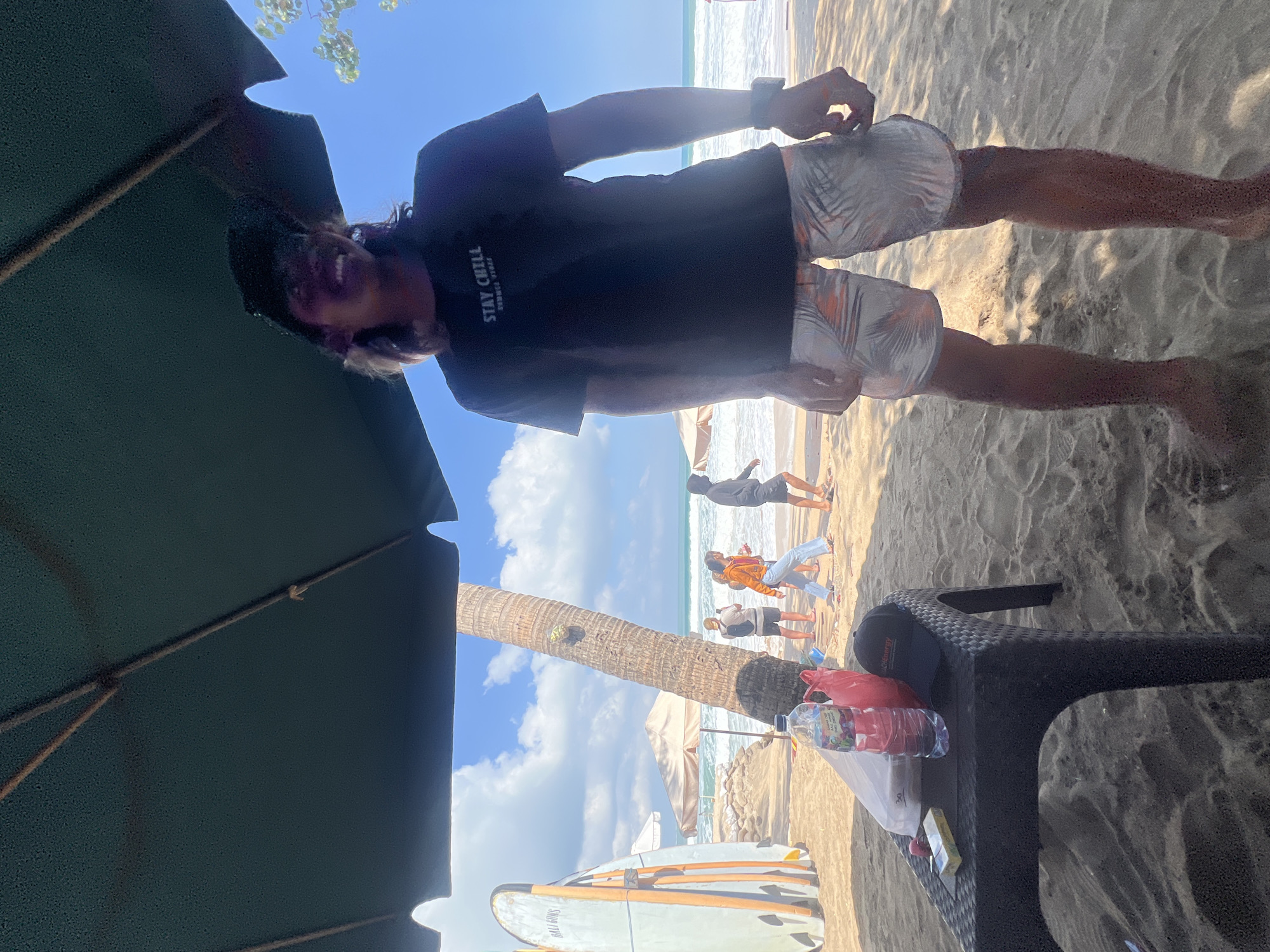 The image shows a man standing on a sandy beach, with a large umbrella providing shade above him. He is wearing a black t-shirt with the text "STAY CHILL SUMMER VIBES" printed on it and patterned shorts. The beach has several surfboards propped up on the left side of the frame, and other people can be seen in the background walking near the water's edge. A black plastic table is in the foreground on the left, with a bottle of water, a red plastic bag, a pack of cigarettes, and a black baseball cap on it. The ocean and a clear blue sky are visible beyond the beach. The setting appears to be Kuta, Indonesia, a popular tourist destination known for its beaches and surf spots.