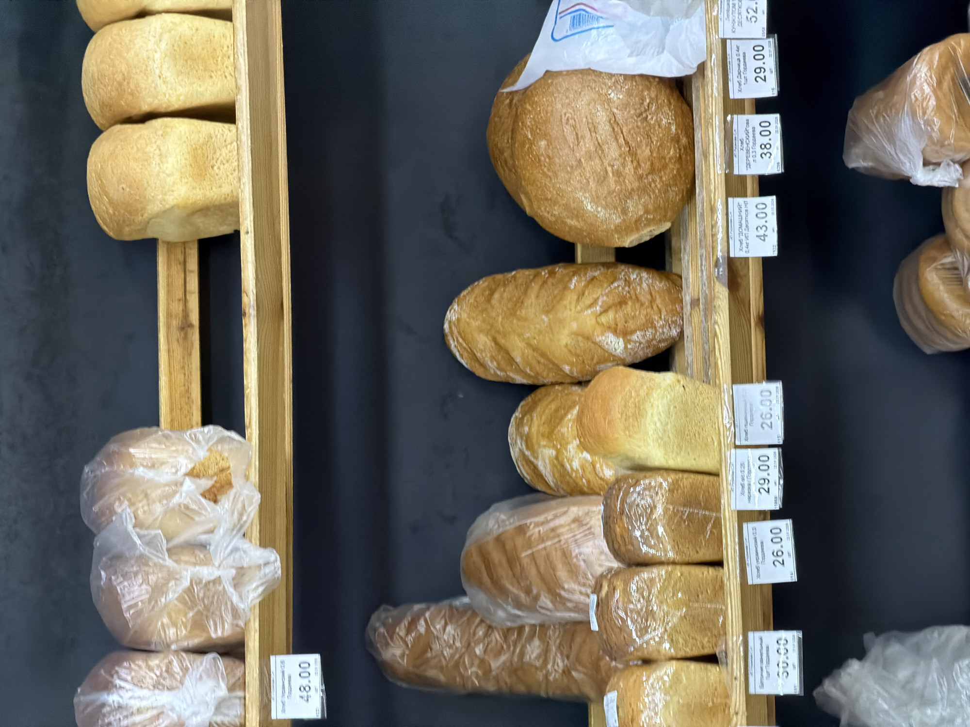 The image shows a shelf of bread loaves in a store, likely a bakery or grocery store. The loaves are of various shapes and sizes, some wrapped in clear plastic. Prices in Russian rubles are displayed on tags next to the loaves. The image was taken in the city of Chaikovsky, Russia, based on the currency displayed on the price tags.