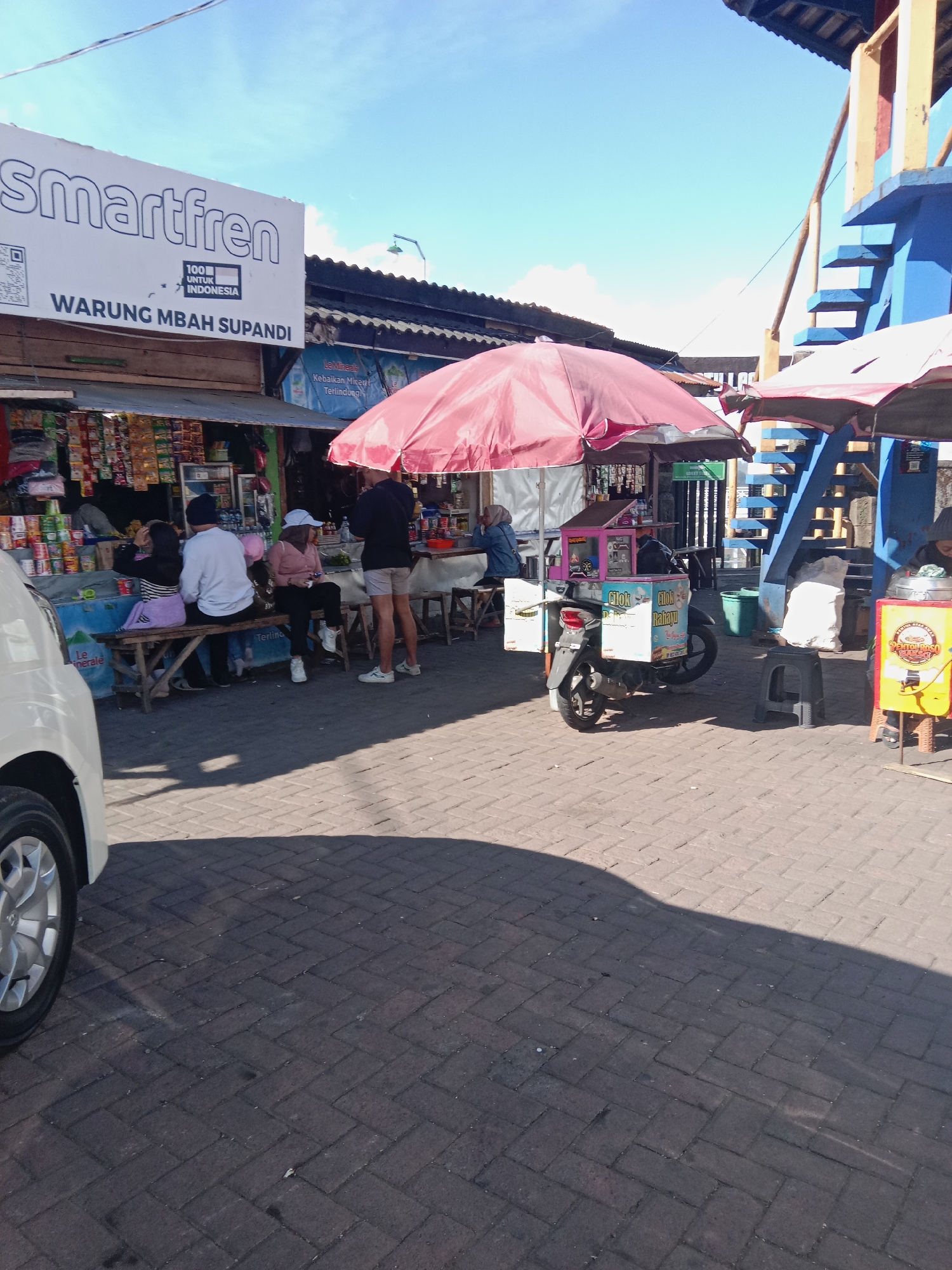 The image depicts an outdoor scene in what appears to be a market or roadside stall area in Tieng, Indonesia. In the foreground, a white car is partially visible on the left. The ground is paved with dark brickwork, with a large shadow cast across it from the car and other structures.
In the mid-ground, several food stalls are set up. A red umbrella shades one area where people are sitting on a wooden bench. One person, wearing a white long-sleeved shirt and a dark beanie, is sitting on the bench. Next to them, a person in a white hat and pink shirt is also seated. Further to the right, a person in shorts and a dark shirt stands near a stall. Another stall features a motorcycle with a colorful cart attached, likely selling food items. This cart has text that appears to read "Cilok Rahayu".
Behind the stalls, a building with a blue and yellow structure and stairs is visible, suggesting a multi-level structure. Signs are present, including one that says "Smartfren" and another with "WARUNG MBAH SUPANDI". Various products are displayed for sale at the stalls, including packaged goods. The sky above is bright blue with some clouds, indicating a sunny day. The overall impression is a lively, commercial street scene.