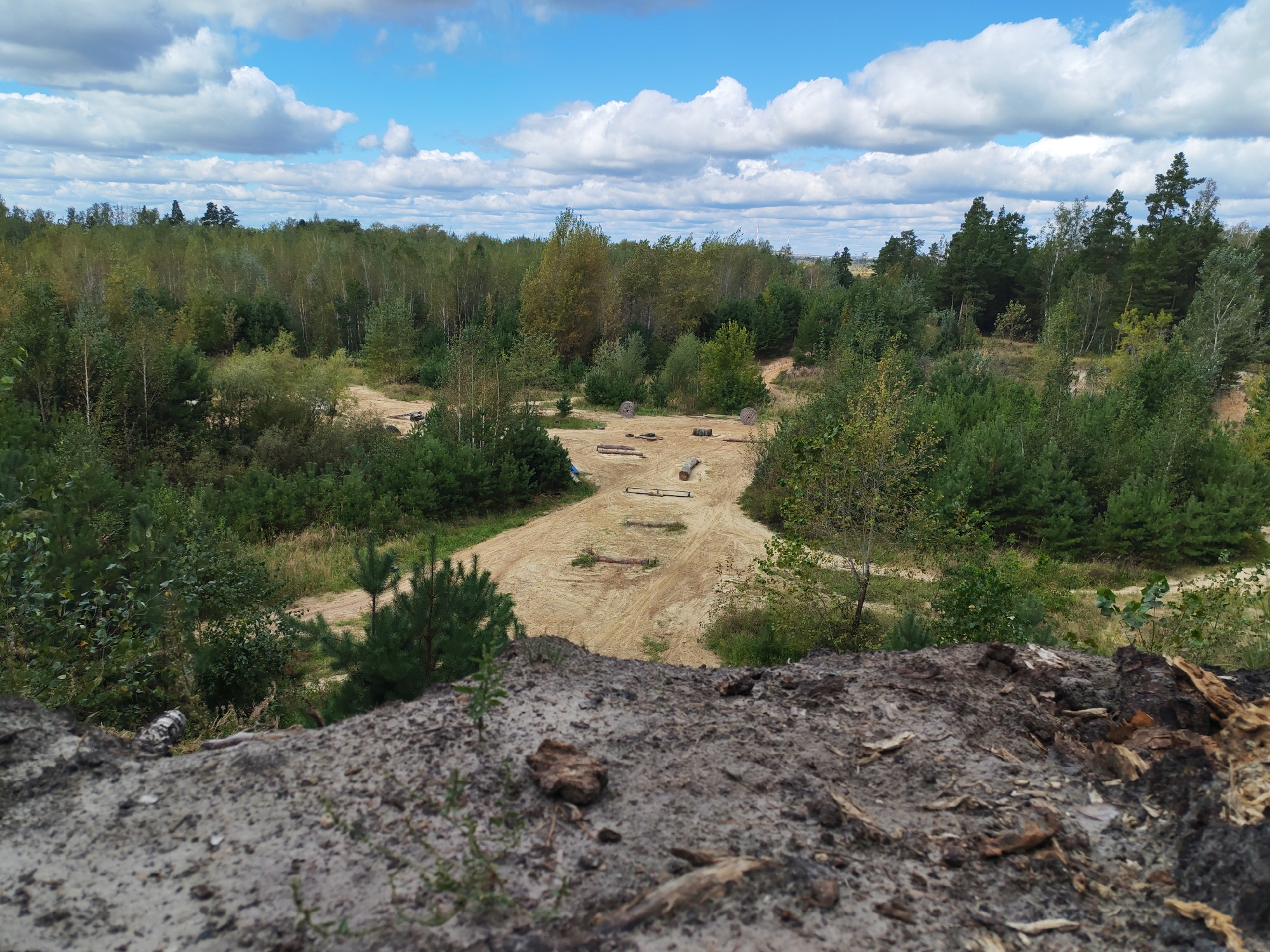 A wide-angle, high-angle outdoor shot captures a sandy track winding through a dense forest under a partly cloudy sky. The track, made of light brown sand, appears to be a constructed obstacle course or path with several logs and what look like tires or barrels placed along it. The forest surrounding the track is a mix of deciduous and coniferous trees, with varying shades of green and some yellowing leaves, suggesting it could be late summer or early autumn. The foreground is dominated by a close-up view of rough, dry dirt and some scattered debris, likely a disturbed patch of ground or a natural embankment. The overall impression is one of a natural or semi-natural environment, potentially used for recreational activities.