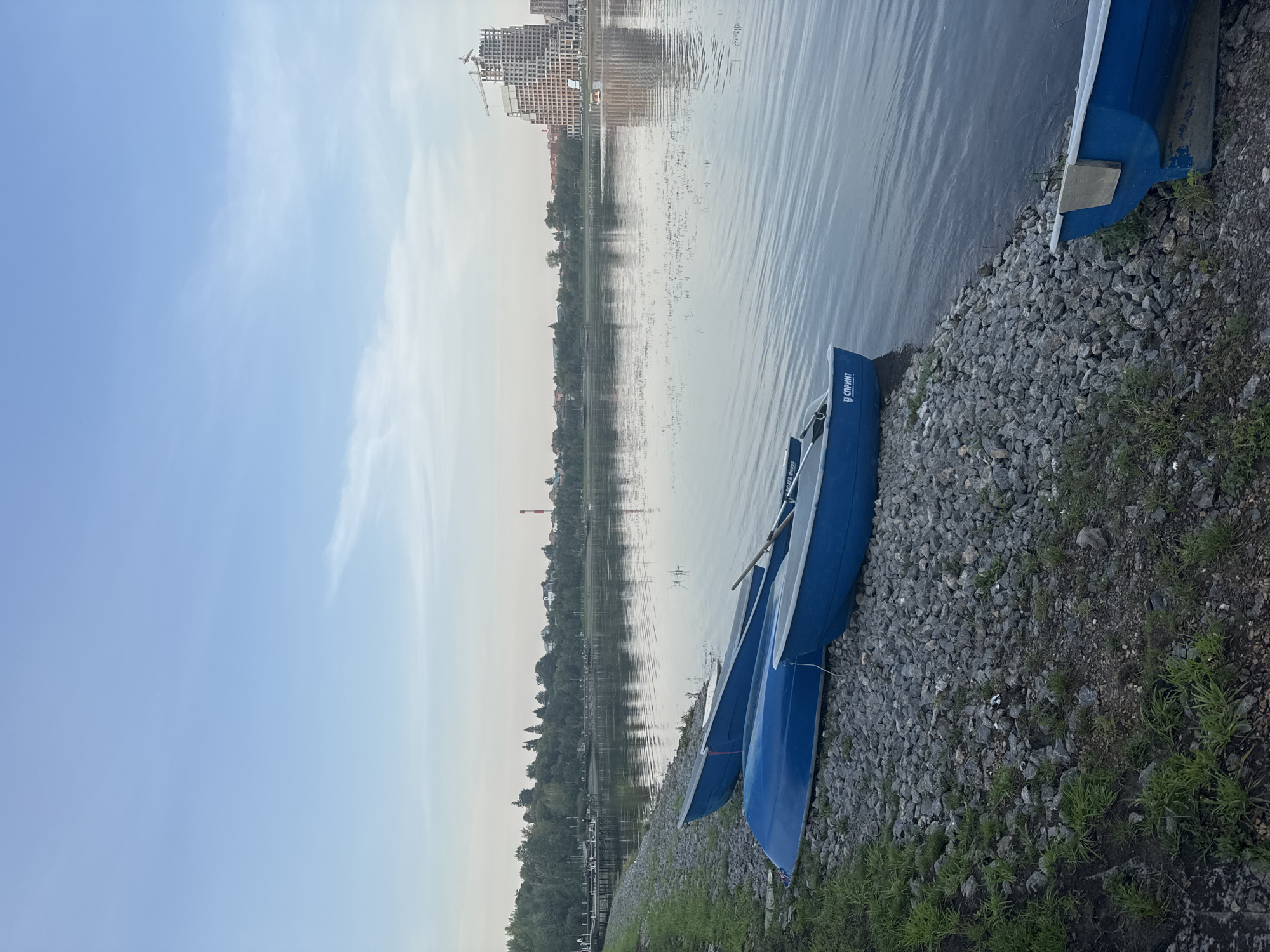 The image captures a tranquil scene along the embankment of the Ufa River in Ufa, Russia. In the foreground, three blue rowboats are pulled ashore, resting on a bed of gray pebbles and sparse green grass. The boats are positioned lengthwise, with two parallel to the water's edge and one slightly angled. One of the boats in the foreground has the Cyrillic word "СПРИНТ" visible on its side.
The Ufa River stretches across the middle ground, its surface rippling gently, reflecting the pale blue sky and the faint silhouettes of trees and buildings on the opposite bank. The water is calm, with occasional patches of aquatic vegetation.
In the background, the city skyline of Ufa is visible. Dominating the right side of the frame is a tall, unfinished building, resembling a skeletal structure of concrete and steel, with construction cranes still present. To the left of this modern structure, a cluster of residential buildings and trees line the riverbank, interspersed with a distant red and white smokestack. A wooden pier extends into the water from the left bank, with a few indistinct figures possibly present on it. The sky is a soft gradient of pale blue, with thin wisps of clouds and a slightly hazier horizon. The overall atmosphere is serene, suggesting either early morning or late evening.