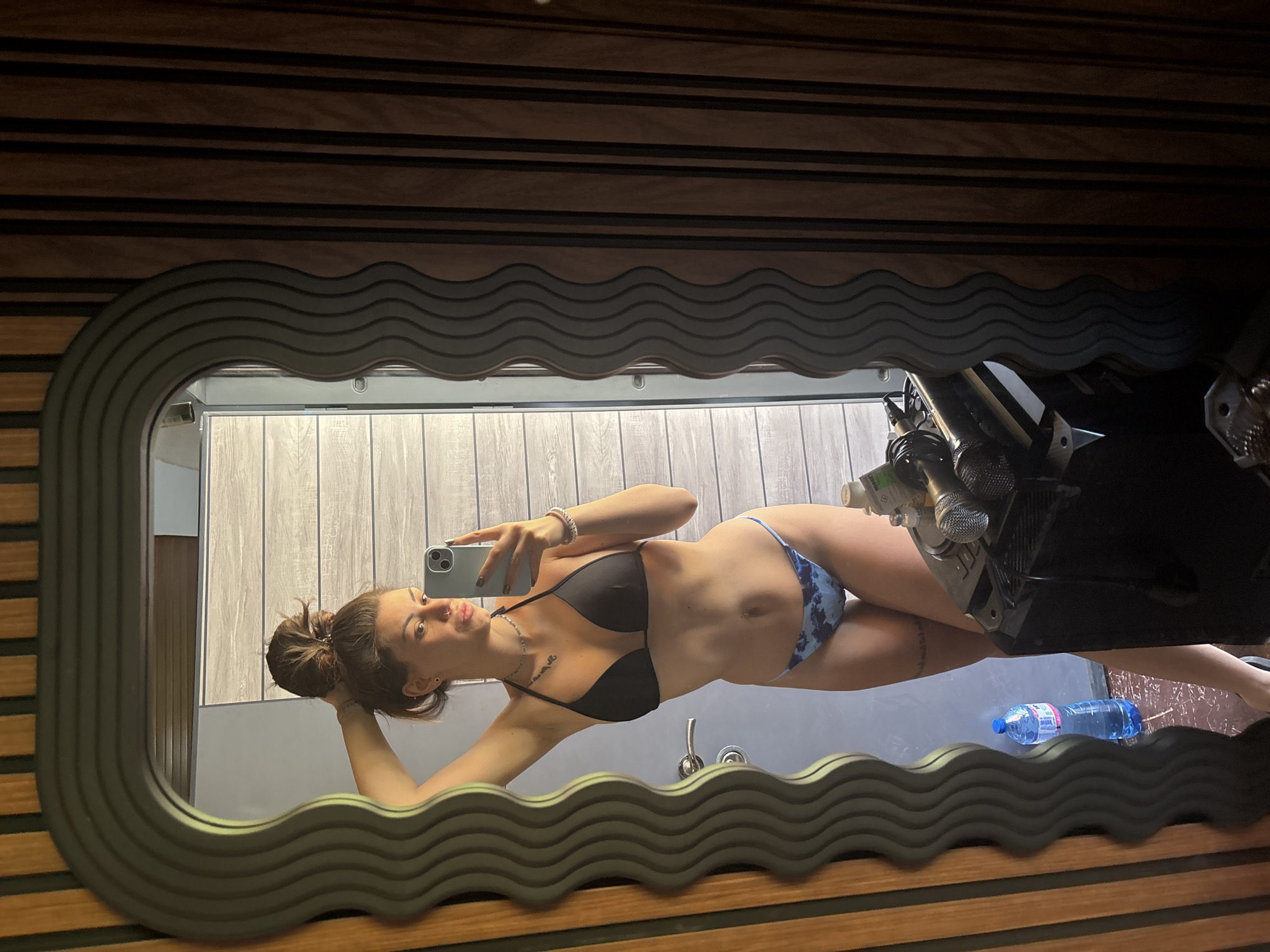 A woman in a black bikini top and blue patterned bikini bottoms is taking a selfie in a mirror. Her hair is in a bun. The mirror is framed by a dark, wavy pattern, and the background appears to be a bathroom with wood-patterned walls. A water bottle is visible near the bottom of the mirror. The photo appears to be taken indoors. No specific details suggest it is in Belopoptsi, Bulgaria.