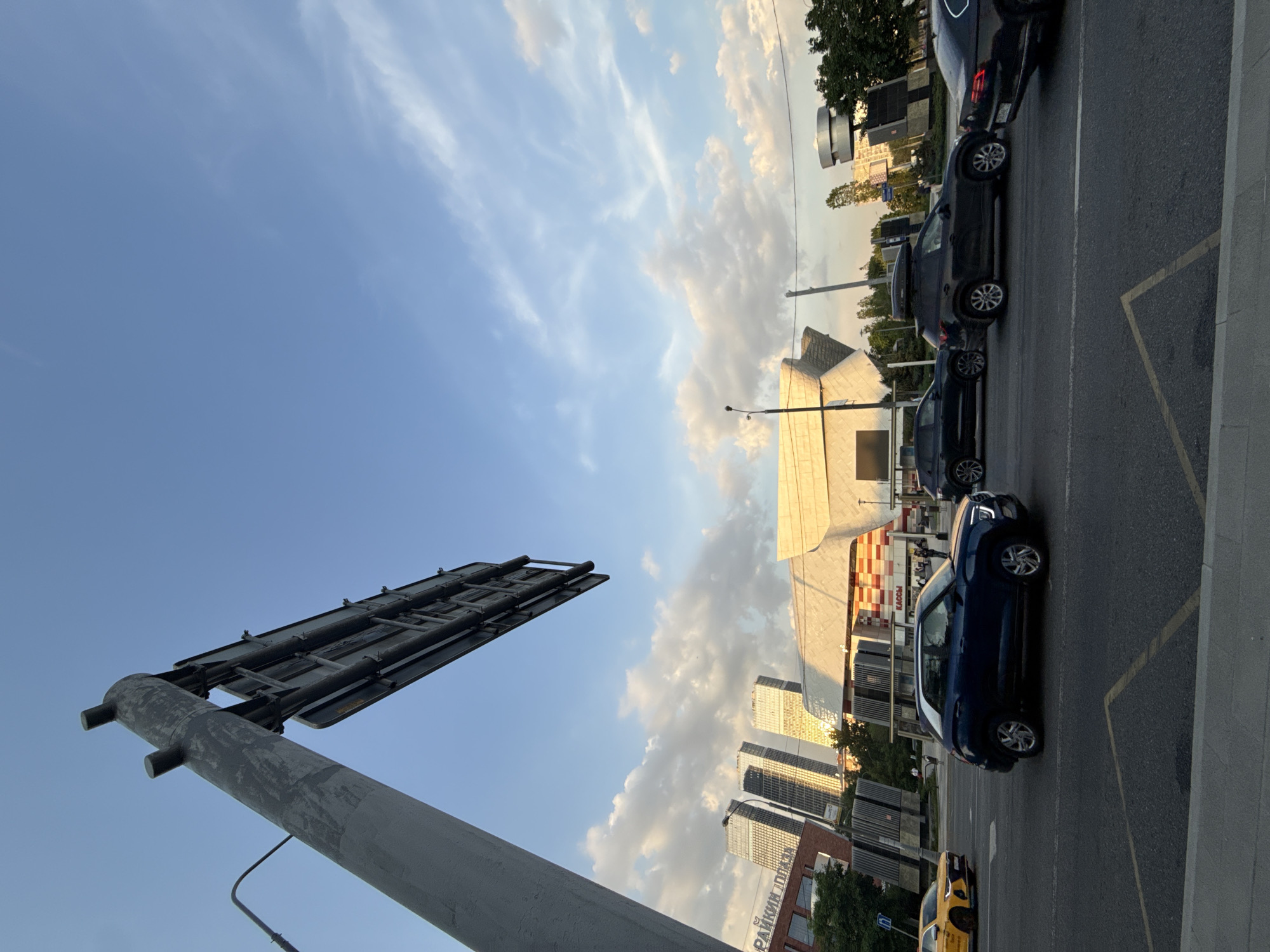 The image captures an urban street scene in Moscow, Russia, under a partly cloudy sky. In the foreground, there is a gray metal pole supporting a large sign structure. A line of vehicles, including a dark blue passenger car, several black SUVs or vans, and a yellow taxi, are parked along the roadside, near yellow painted lines. Several buildings are visible in the background. The buildings are modern and some have reflective glass facades. Text in Cyrillic can be seen on a sign on one of the buildings.
