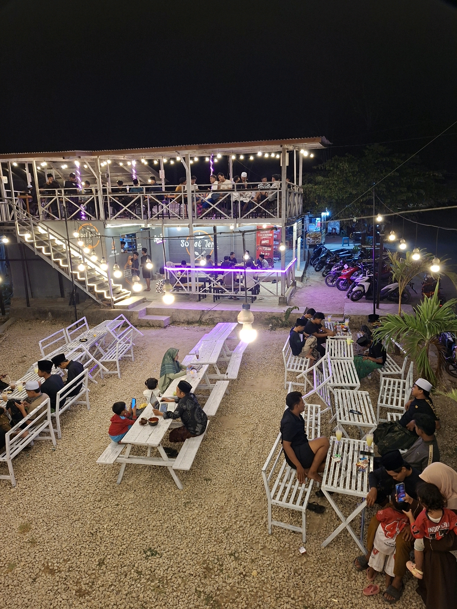 The image depicts an outdoor dining area at night, likely a restaurant or cafe, situated in Lantek Barat, Indonesia. The establishment features a multi-level structure with an upper deck illuminated by strings of lights and purple LED accents. Several people are visible on the upper deck, seated at tables and interacting. Below, the ground is covered in gravel and features numerous white picnic tables and benches.
In the lower portion of the image, groups of people are gathered at the tables. One table has individuals, including a child, seated with a laptop. Another group is seated at a table with drinks. To the right, a family, including two young children, is present, with one child being held. In the background, a row of motorcycles is parked, and the sky is dark. The overall scene suggests a lively, social gathering space in the evening.