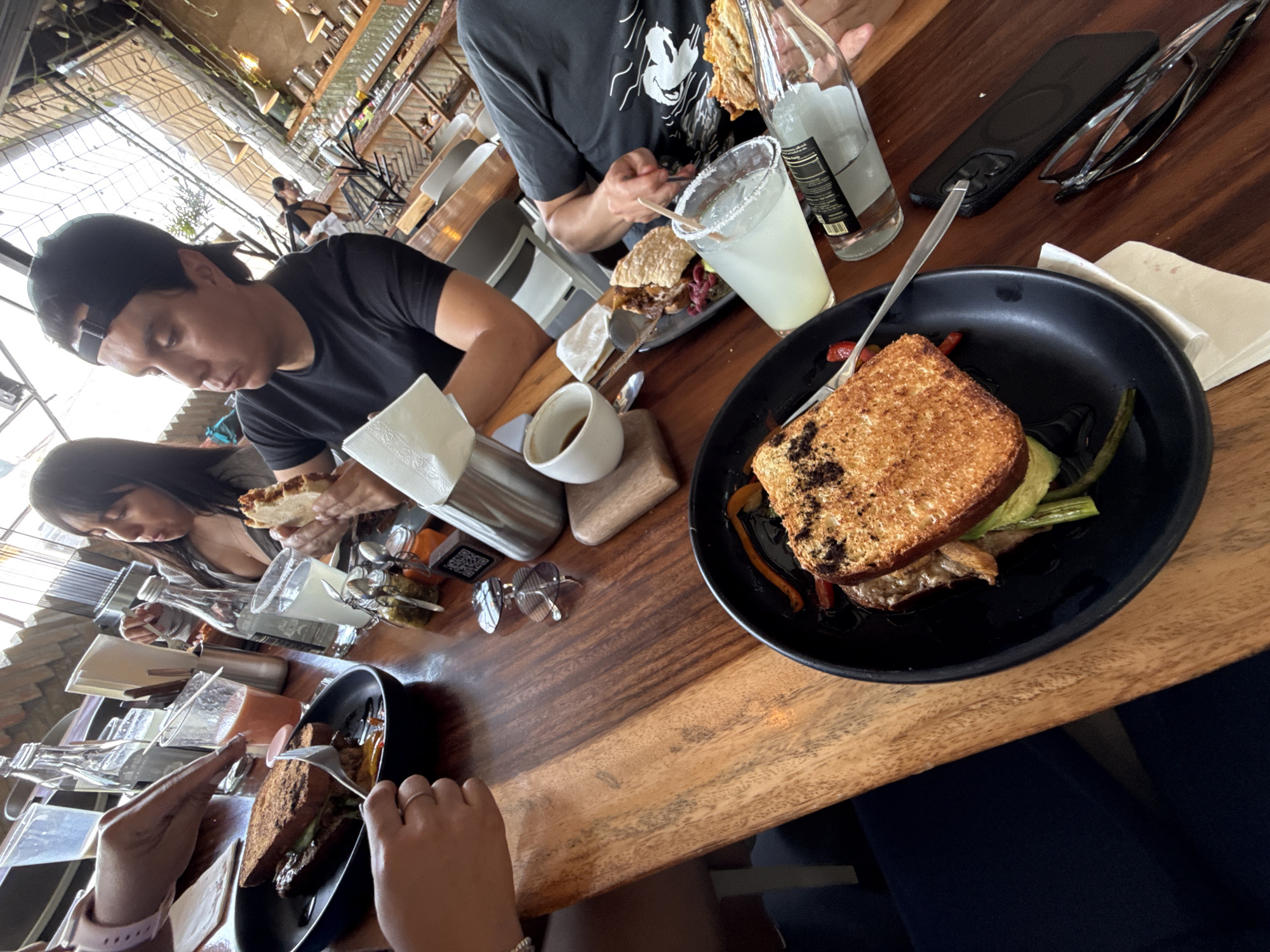 A restaurant scene shows three people eating at a wooden table.
A man in a black t-shirt and baseball cap is on the left side of the table. A woman with dark hair is next to him, also eating. Across from them is a man in a black t-shirt with a Mickey Mouse graphic. One diner wears a pink watch on the left-hand side of the frame.
Food and drinks are placed on the table. A black plate holds a sandwich with asparagus and peppers. There is a glass of a pale-colored beverage and a bottle of clear liquid.
Glasses and silverware are scattered around the table. A black cell phone and glasses are also on the table. The restaurant has a casual atmosphere with visible windows.
The location is San Andrés Cholula, Mexico.