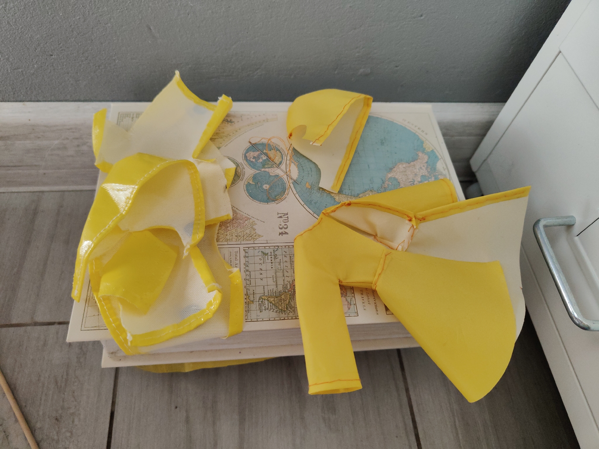 The image displays a close-up, overhead view of a stack of books topped with yellow fabric pieces. One piece of yellow fabric is shaped like a stylized leg or arm, attached to a book. Another piece, also yellow, is draped over the books, appearing torn or unfinished, with some white fabric visible. A needle and orange thread are present on the map section of the top book, suggesting ongoing craft work. The books are open, revealing maps and text. The background consists of a gray wall, a white cabinet with a silver handle, and a tiled floor. The scene is indoors, on what appears to be a domestic setting. There is no indication of the city of Wrocław, Poland in the image.