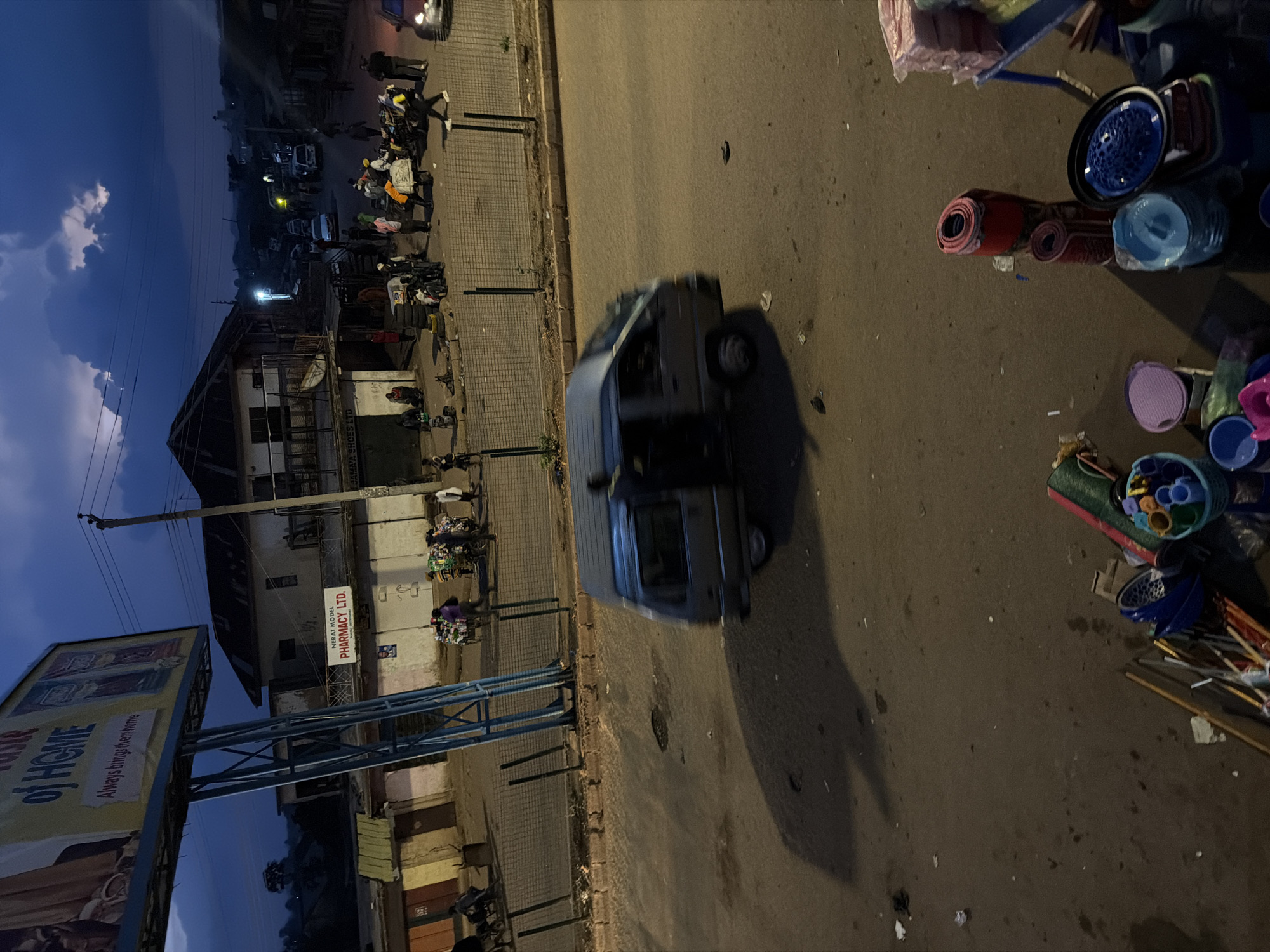 The image captures a street scene in Bukuru, Nigeria, likely during dusk or early evening. In the foreground, a grey van is blurred due to motion, indicating it is moving. To the right, a small stall displays various household items including rolled mats, plastic bowls, and buckets. The background reveals a bustling street with buildings, a billboard advertising "OF HOME," and a sign for "NERAT MODEL PHARMACY LTD." Several people are visible, some walking, others gathered in small groups, and a few are seen pushing carts laden with goods. The overall environment suggests a commercial area with active pedestrian and vehicular traffic. The sky is dark blue with some clouds, and streetlights illuminate parts of the scene.