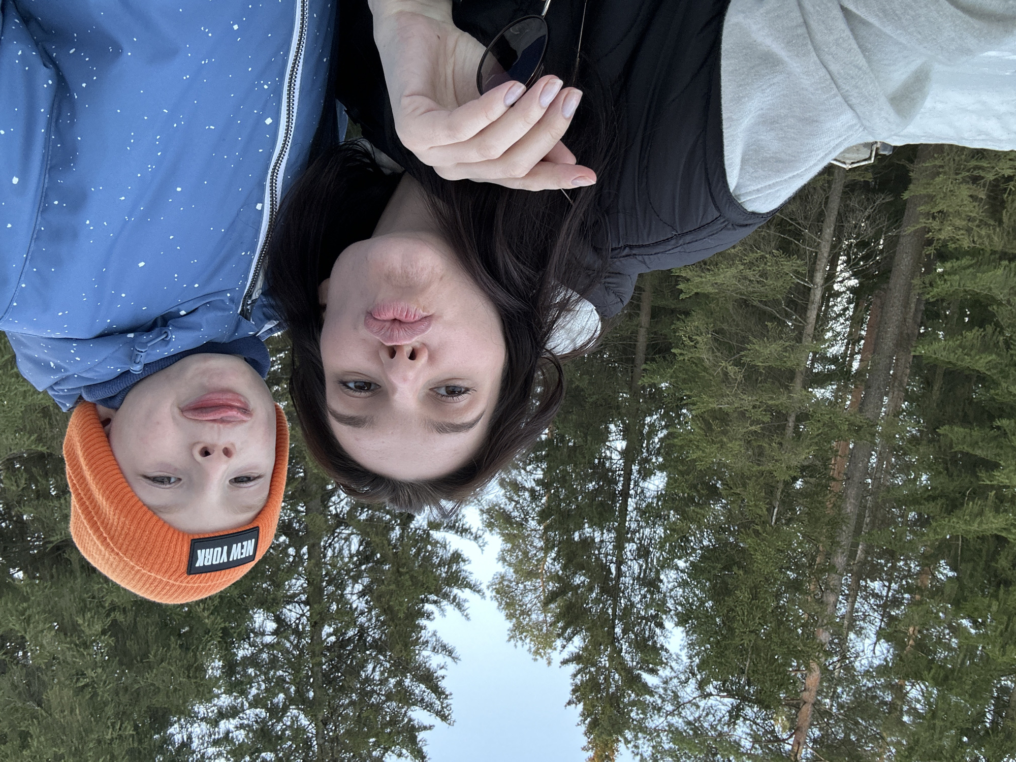 The image depicts an outdoor scene in a coniferous forest in Прикамский, Russia, featuring two individuals posing for a close-up photograph. An adult female, positioned on the left, has dark hair, light eyes, and is wearing a black puffer jacket over a light grey long-sleeved garment. She is making a "kissing" face with pursed lips and holds dark-lensed sunglasses in her right hand. To her right is a young male child, wearing an orange knit beanie with a black "NEW YORK" patch and a blue hooded jacket adorned with white speckles. The child is looking towards the camera with his tongue slightly extended. The background consists of numerous tall, green pine or fir trees, with visible patches of snow on the ground in the lower-left corner. A light blue sky is visible through the tree canopy.