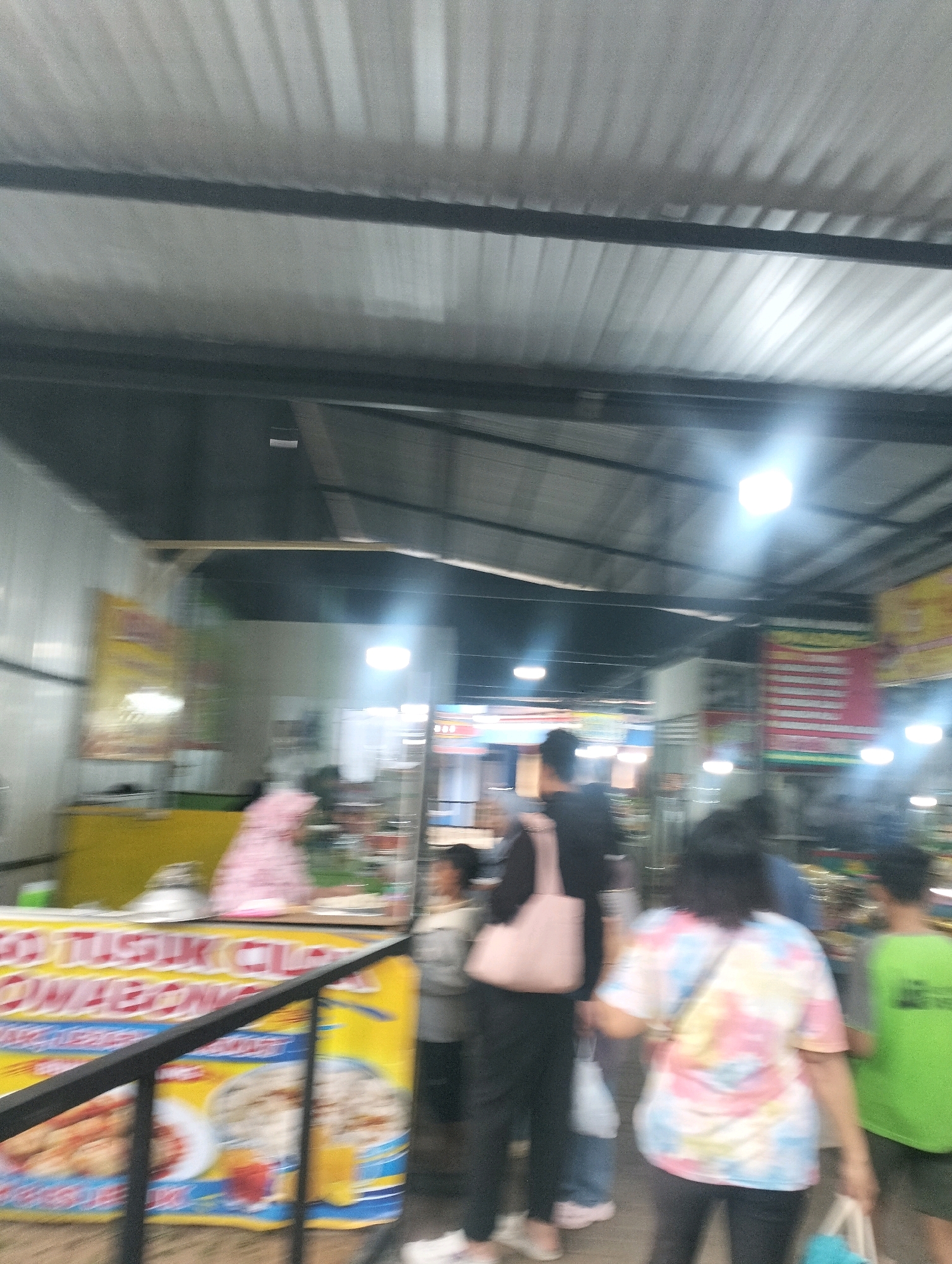 The image depicts an outdoor market or food stall area, likely in Karangbanjar, Indonesia. The scene is characterized by overhead structures with corrugated metal roofing, casting striped shadows. Several individuals are visible, some walking and others interacting with the stalls. In the foreground, a yellow banner with Indonesian text and images of food, including what appears to be fried items and bowls of rice, is partially obscured by a black railing. The banner's text includes "CO TUSIK CUCUMBONG." A person in a pink patterned dress stands behind the banner. Further into the scene, other individuals are seen with shopping bags and wearing casual attire, including a person in a tie-dye t-shirt. Lighting appears to be a mix of overhead fluorescent lights and natural light, contributing to a somewhat blurred and hazy overall impression. The environment suggests a bustling commercial area with food vendors.
