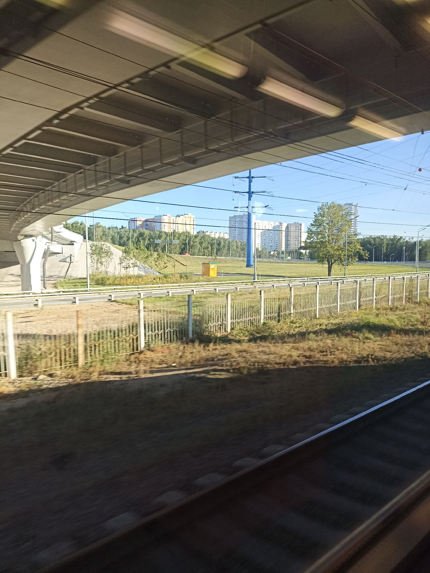 The image depicts a view from a train window, looking out at an urban landscape under a clear blue sky. In the foreground, train tracks are visible, with a blurred effect suggesting movement. Above the tracks, a large concrete overpass or bridge structure dominates the upper portion of the frame, its underside illuminated by artificial lights. Beyond the overpass, a wide, grassy field stretches out, dotted with trees and a lone, mature tree standing in the middle. A white metal fence runs along the edge of the field, separating it from a road or pathway. In the distance, several multi-story apartment buildings are visible, characteristic of urban residential areas. A tall, blue electricity pylon stands prominently in the mid-ground, with power lines crisscrossing the sky. A small, yellow utility box is situated near the fence in the mid-ground. The scene appears to be in a modern part of Moscow, Russia, likely a suburban area given the combination of infrastructure and green space. There are no people or animals visible in the frame.