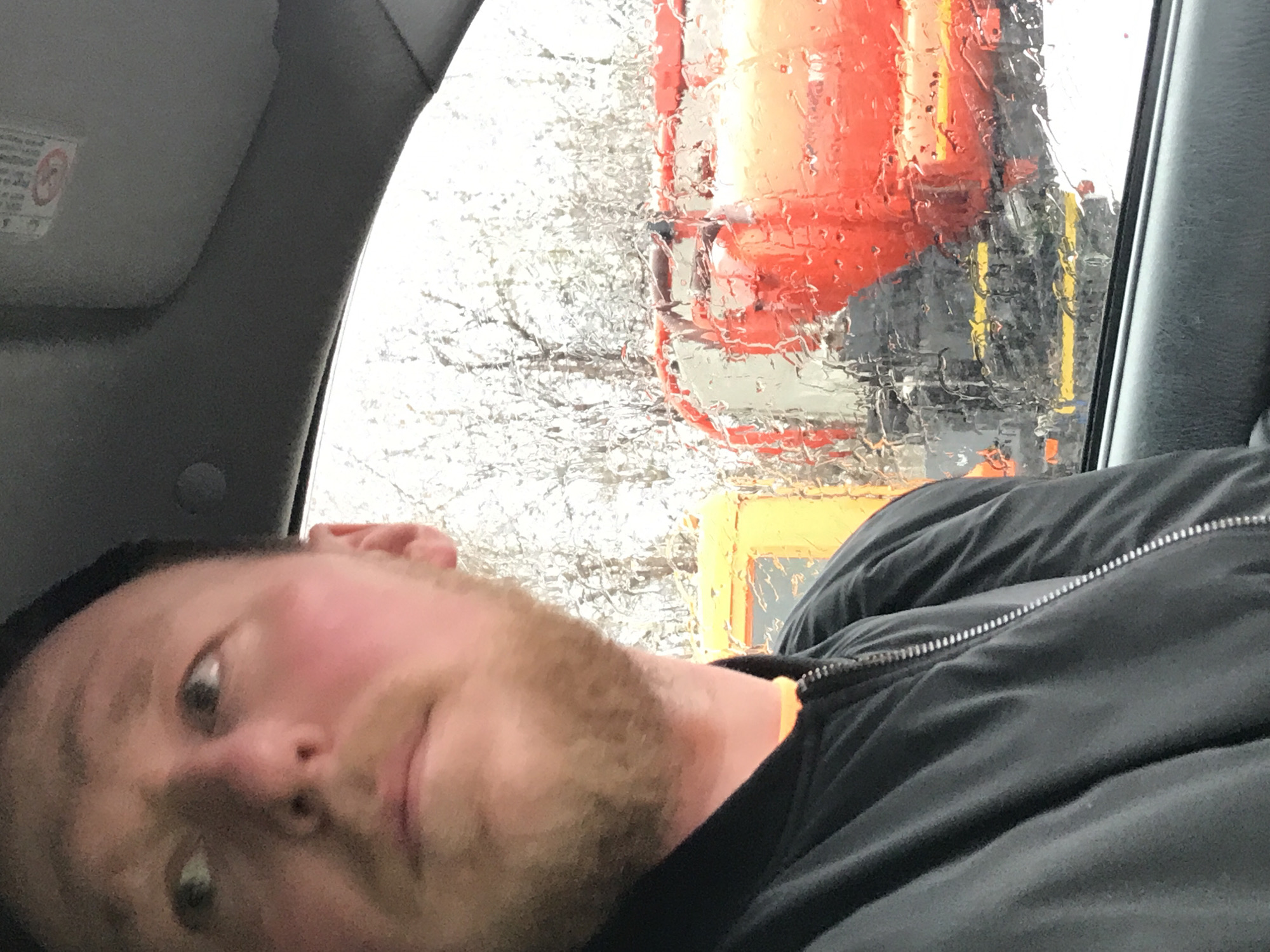 A selfie of a man taken inside a vehicle, presumably in Perm, Russia, given the presence of Russian construction equipment visible through the rain-streaked window. The man has fair skin, a light beard, and is wearing a black zip-up jacket. Outside, visible through the window, is what appears to be a construction vehicle, primarily orange with black and yellow accents, blurred by raindrops. Bare tree branches are also visible behind the vehicle. The vehicle's interior is visible, with elements like the headliner and window frame in view. The photo is taken at an angle.