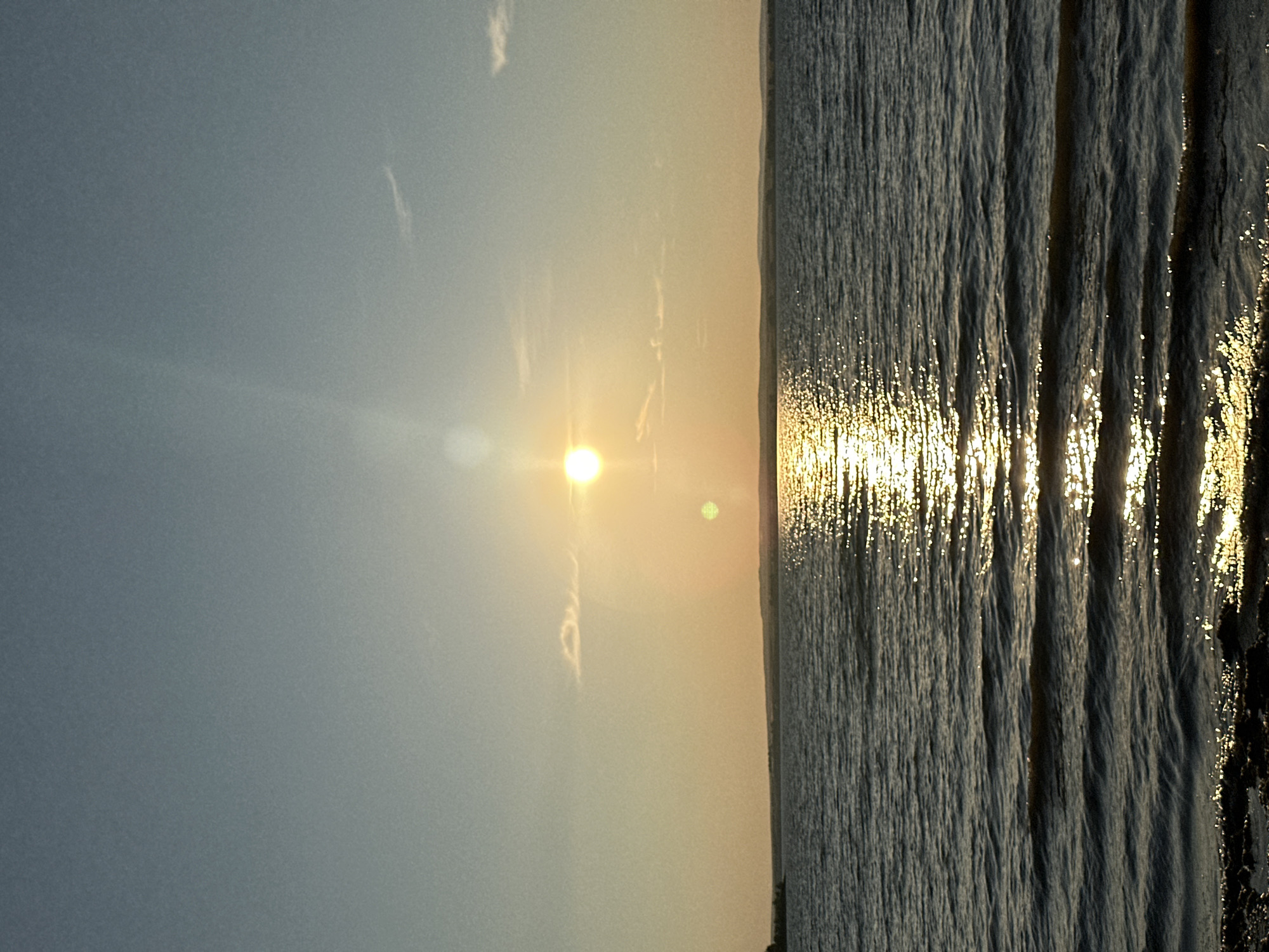 The image depicts a serene sunset over a body of water, likely a lake or reservoir, with a faint shoreline visible in the distance. The sun, a bright orb, is positioned in the upper half of the frame, casting a warm, golden glow across the scene and creating a shimmering reflection on the water's surface. The sky transitions from a clear, pale blue at the top to a softer, yellowish hue near the horizon. A few wispy clouds are present, subtly catching the sunlight. The water itself is calm, with gentle ripples creating textured patterns that catch the light. The foreground shows a rocky shoreline, suggesting the vantage point is from the edge of the water. The overall impression is one of tranquility and natural beauty. This scene could potentially be located near the city of Energetik, Russia, which is situated near Lake Baikal, a vast freshwater lake that experiences spectacular sunsets.