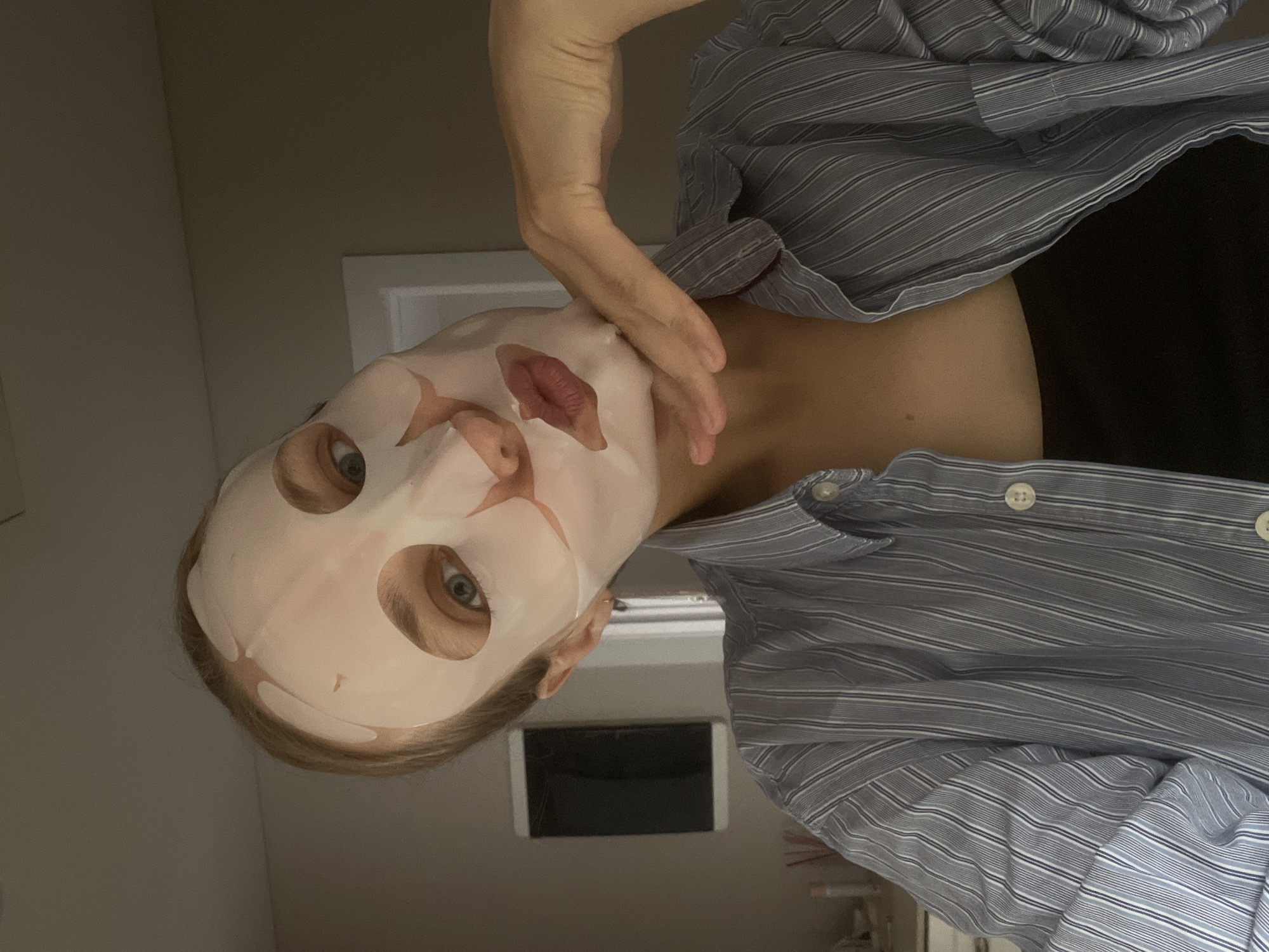A person wearing a white face mask is visible, appearing to be adjusting the mask with their hand. The person is wearing a striped shirt. The background is blurry, but there is a doorway visible. The location is undetermined.