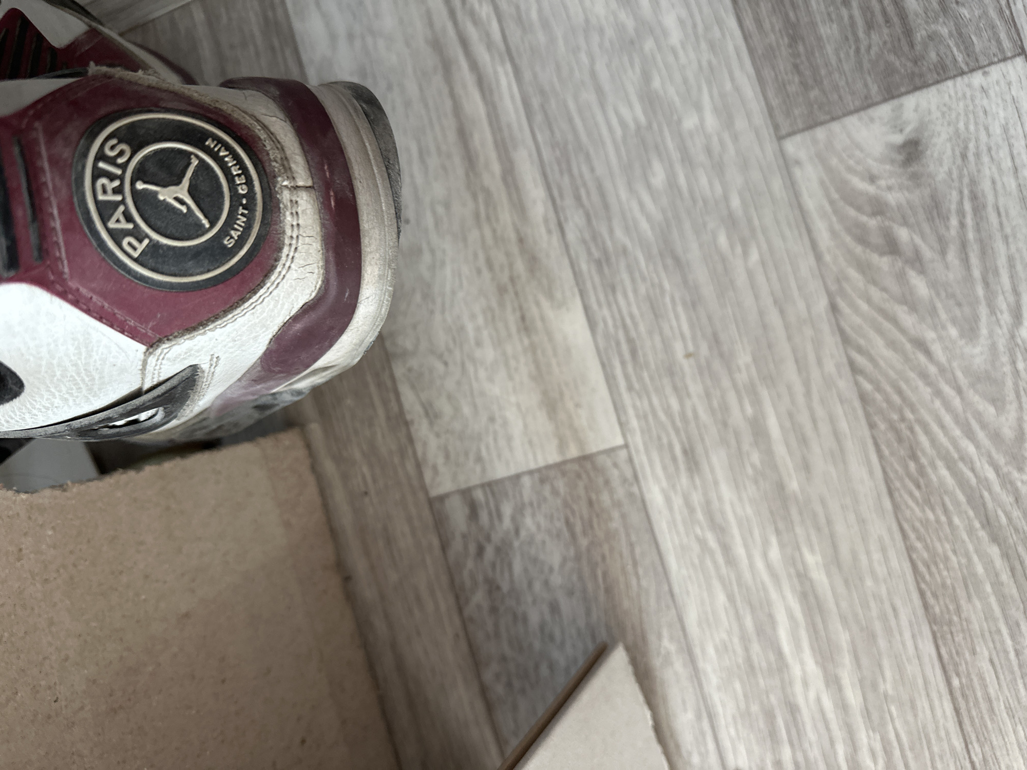 The image shows the heel of a white and maroon Air Jordan sneaker with a Paris Saint-Germain logo, resting on a light-colored wooden floor. A portion of a cardboard box is visible in the upper left corner. The scene is indoors, and there is no indication of the specific location within Birobidzhan, Russia, or any people or actions taking place.