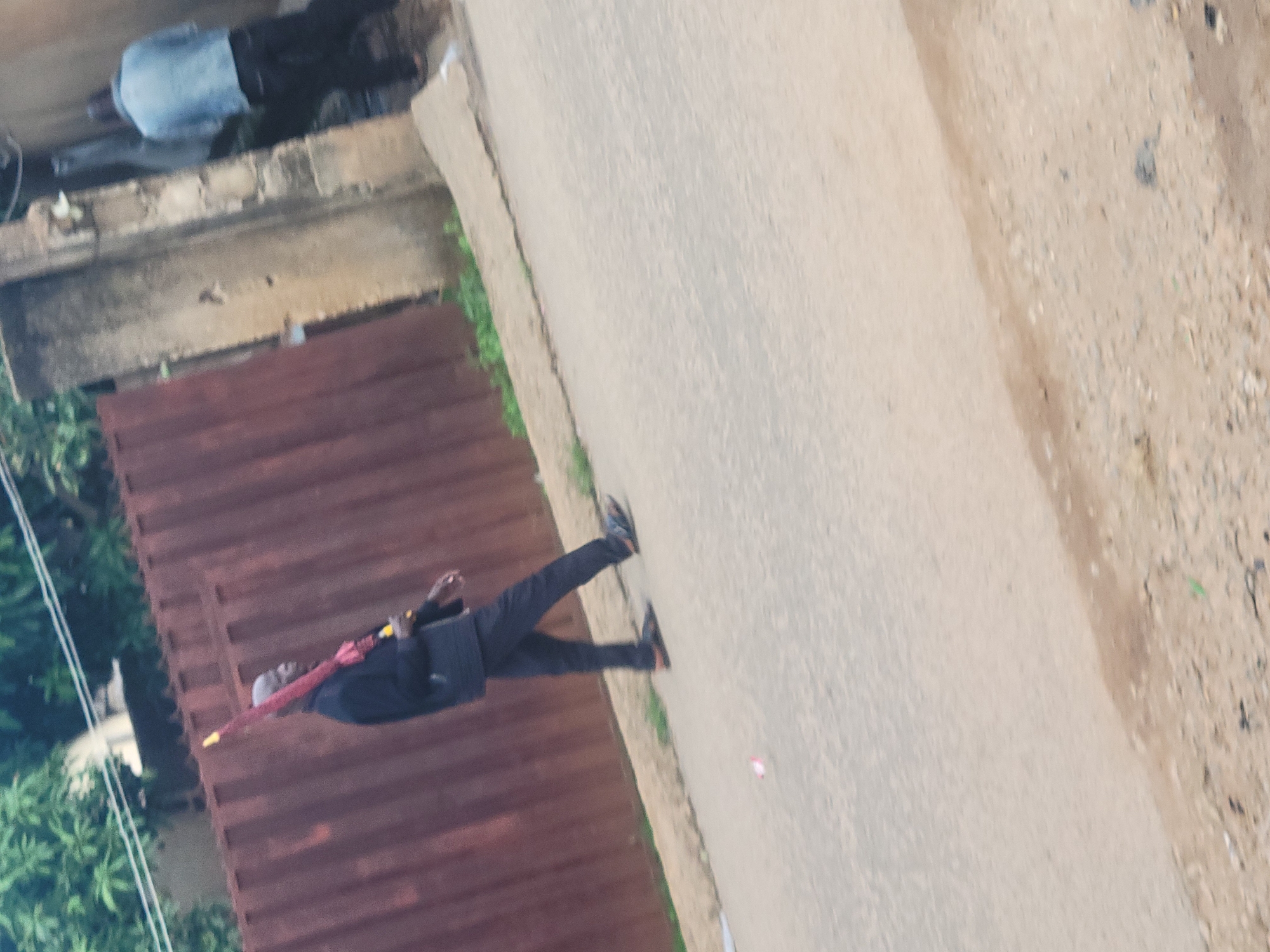This image depicts a street scene in Jos, Nigeria, showing two adult males and environmental features. In the foreground, a bald, dark-skinned male walks left-to-right, facing left. He wears a dark long-sleeved shirt or jacket, dark trousers, and dark sandals with brown accents. He carries a closed red umbrella with a yellow tip over his right shoulder. Behind him, towards the right, another male is partially visible from behind, wearing a light blue jacket and dark trousers. He appears to be positioned near a weathered concrete pillar. The background features a rusty brown corrugated metal fence extending horizontally across the frame, with green foliage visible above it on the far left. The ground is a light grey/beige paved road, appearing somewhat rough with scattered dirt and small stones. A narrow strip of green weeds and a slight curb separate the road from the fence line. A small, reddish-pink piece of litter rests on the road surface in the mid-left foreground. The scene is illuminated by daylight.