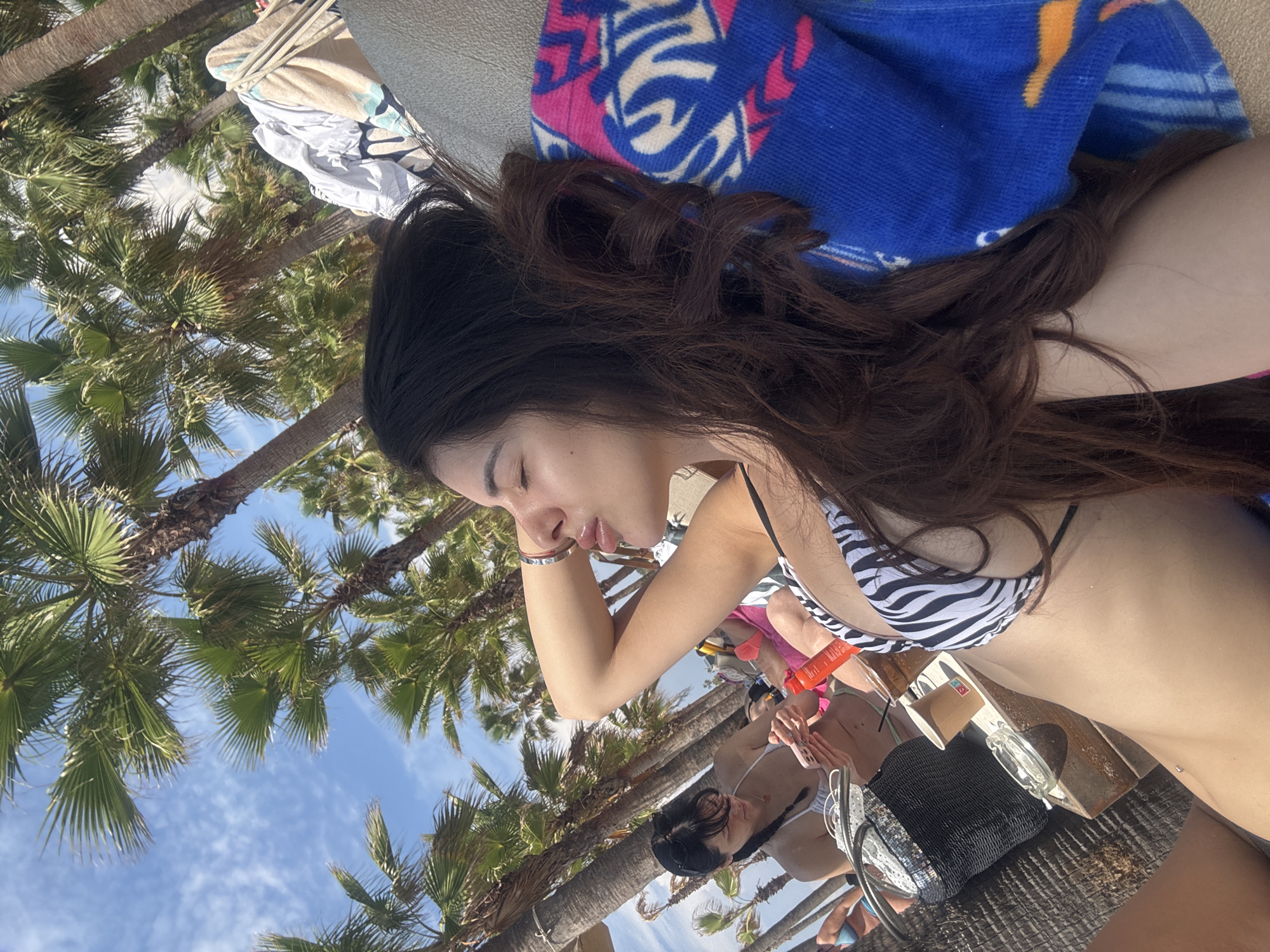 This is a medium shot of two women lounging outdoors, possibly at a beach resort in Keramoti, Greece. The primary focus is on a woman in the foreground, lying on her side with her eyes closed and her head tilted back, her right arm bent behind her head. She is wearing a black and white zebra-print bikini top. Her long, dark hair is spread out around her head and shoulders.
To her left and slightly behind, another woman is seated, looking down at her phone. She is wearing a white bikini top and her hair is pulled back in a braid. They are both positioned on lounge chairs, with a table between them holding a black straw bag, a disposable cup, and an ashtray. A bright orange bottle, likely sunscreen, is also visible on the table. A blue towel with a colorful pattern is draped over the back of the lounge chair in the foreground.
The background is dominated by palm trees, suggesting a warm, tropical or Mediterranean climate. The sky above is a bright blue with scattered white clouds. The overall impression is one of relaxation and leisure in a sunny, outdoor setting.