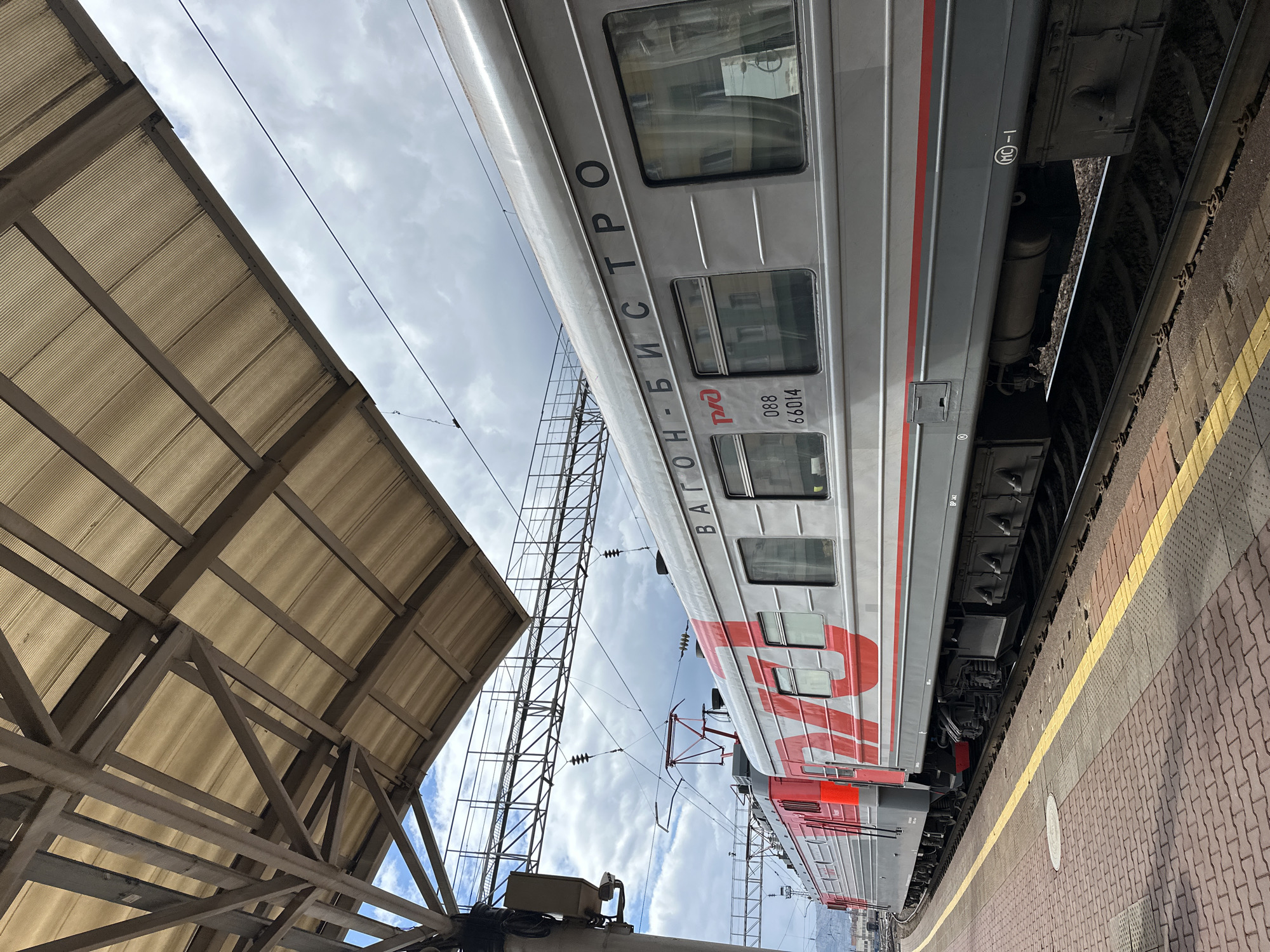 The image shows a train stopped at a station. The train is silver and red and has the words "ВАГОН-БИСТРО" written on the side, which translates to "Restaurant Car". The train car also displays the number "088 66014". The train is next to a platform with a yellow line. Overhead, there is a metal structure providing shelter, with a cloudy sky visible above. The location of the train and station is in Krasnoyarsk, Russia.