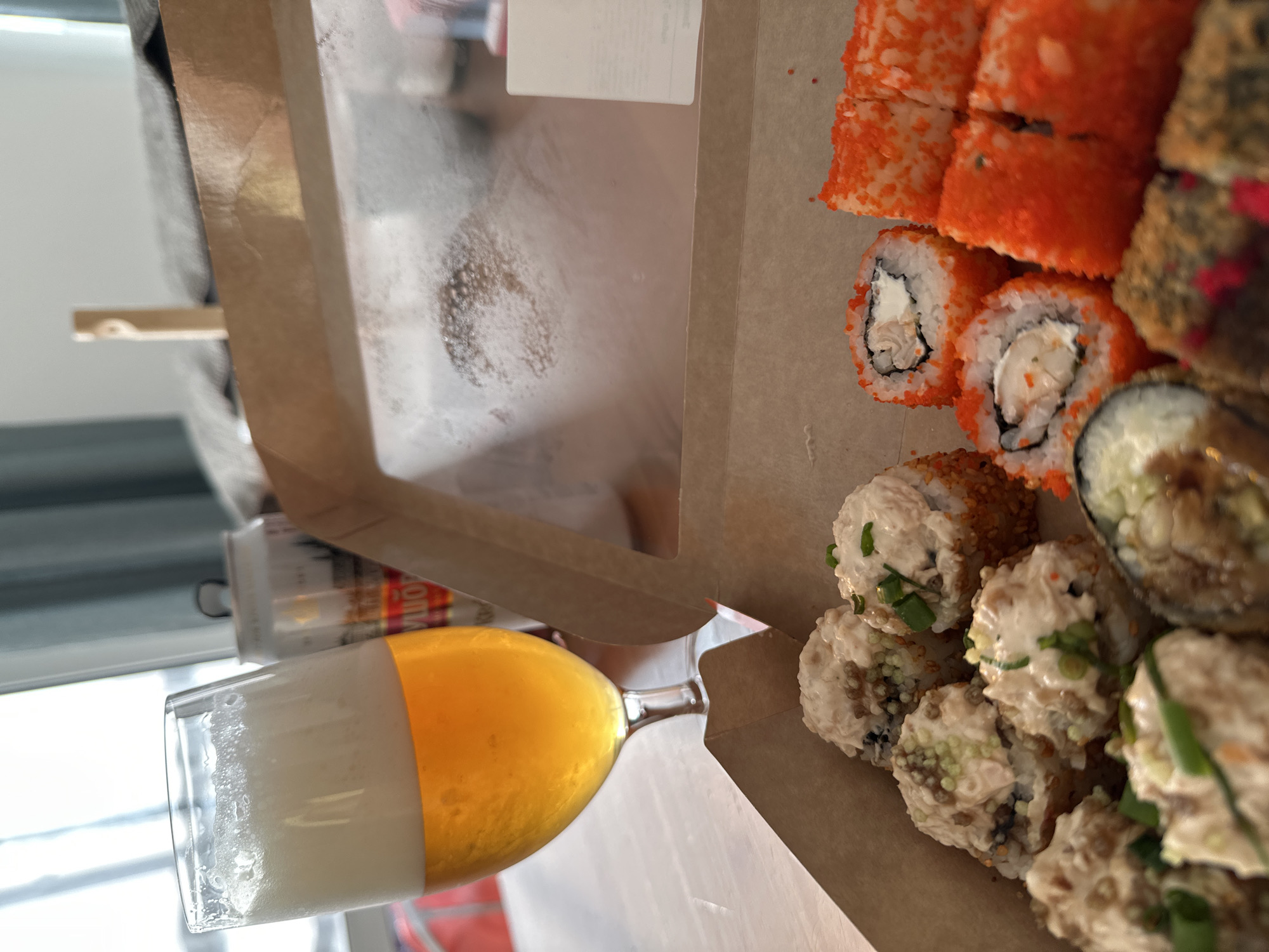 A glass of beer and a box of sushi sit on a table. The beer is a pale yellow color with a foamy head. The sushi is arranged in a brown cardboard box and includes several different types of rolls, some covered in orange roe. In the background, a can of beer and a blurry window are visible. The overall scene suggests a casual meal.