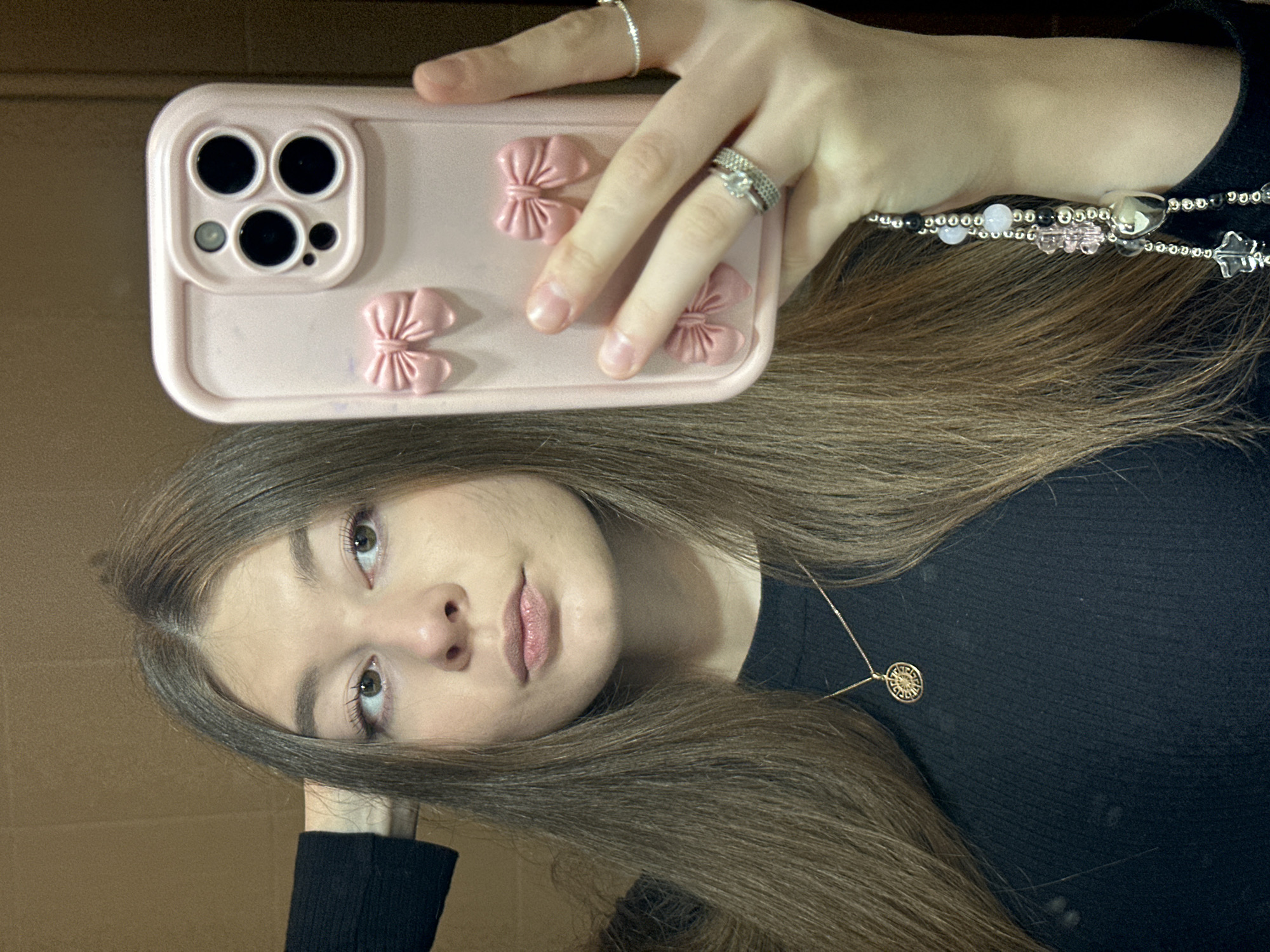 A young woman is depicted in a mirror selfie, holding a light pink smartphone. The image is rotated 90 degrees counter-clockwise. Her left hand, adorned with multiple silver-toned rings (a simple band on the index finger and two stacked rings, one with stones, on the ring finger), holds the phone. The phone case features a triple-lens camera module and three decorative pink bows. A beaded strap or lanyard, composed of silver, black, and light pink opaque beads, along with clear heart and leaf-shaped beads, is visible around her wrist or hand. The woman has long, straight, medium-brown hair, parted down the middle, and light-colored eyes. She wears a black ribbed top and a delicate gold-toned necklace with a circular, patterned pendant. Her expression is neutral and direct towards the camera. The background is a plain, light brown wall or surface, indicating an indoor setting. No text is visible. The location context for this scene is Kazan, Russia.