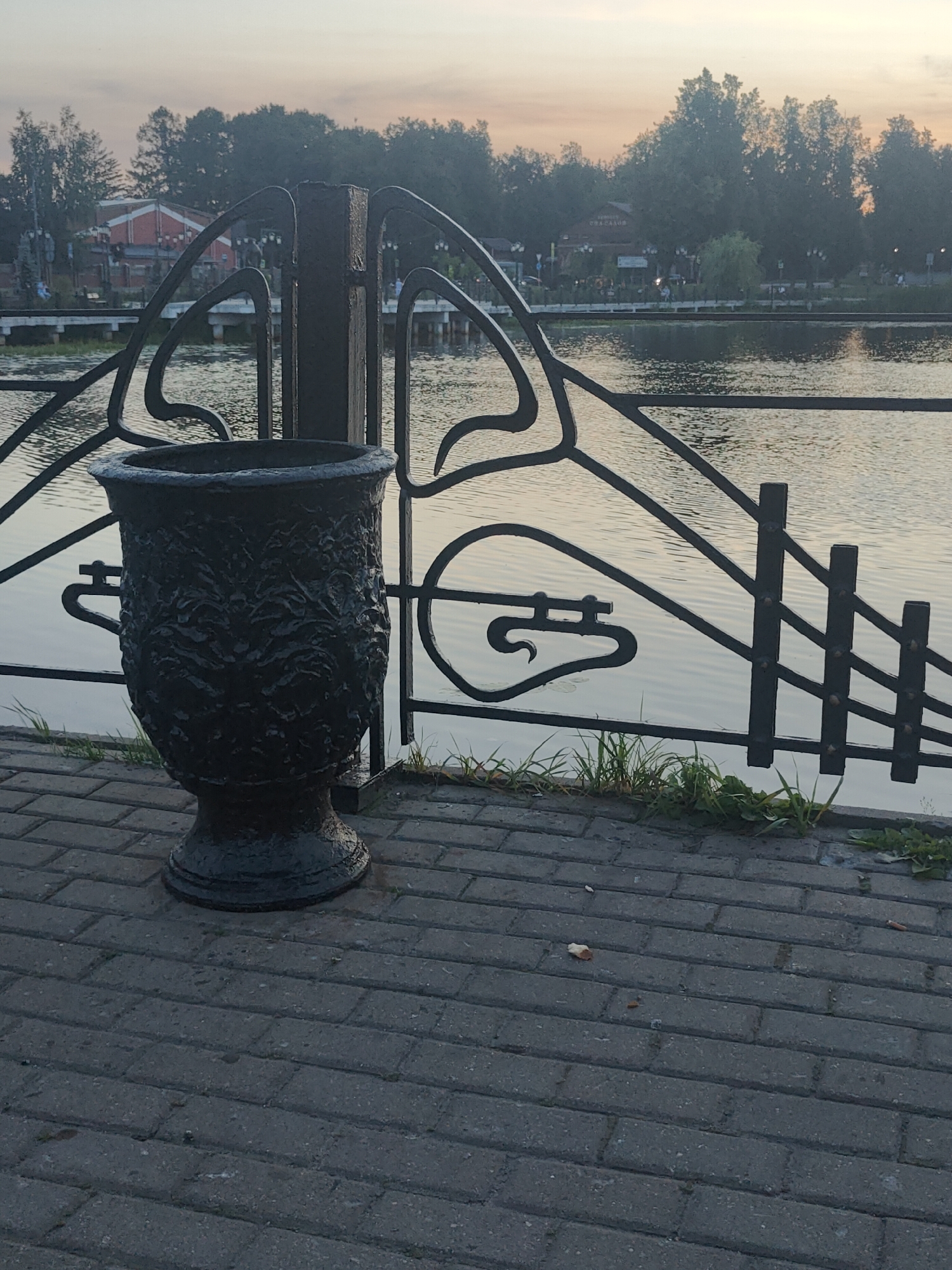 A black ornamental trash receptacle stands on a cobblestone pathway next to a black metal railing. The railing separates the pathway from a body of water. Trees and buildings are visible across the water. The sky is a soft orange and grey, suggesting either sunrise or sunset. The scene appears to be an outdoor public space in a city.
