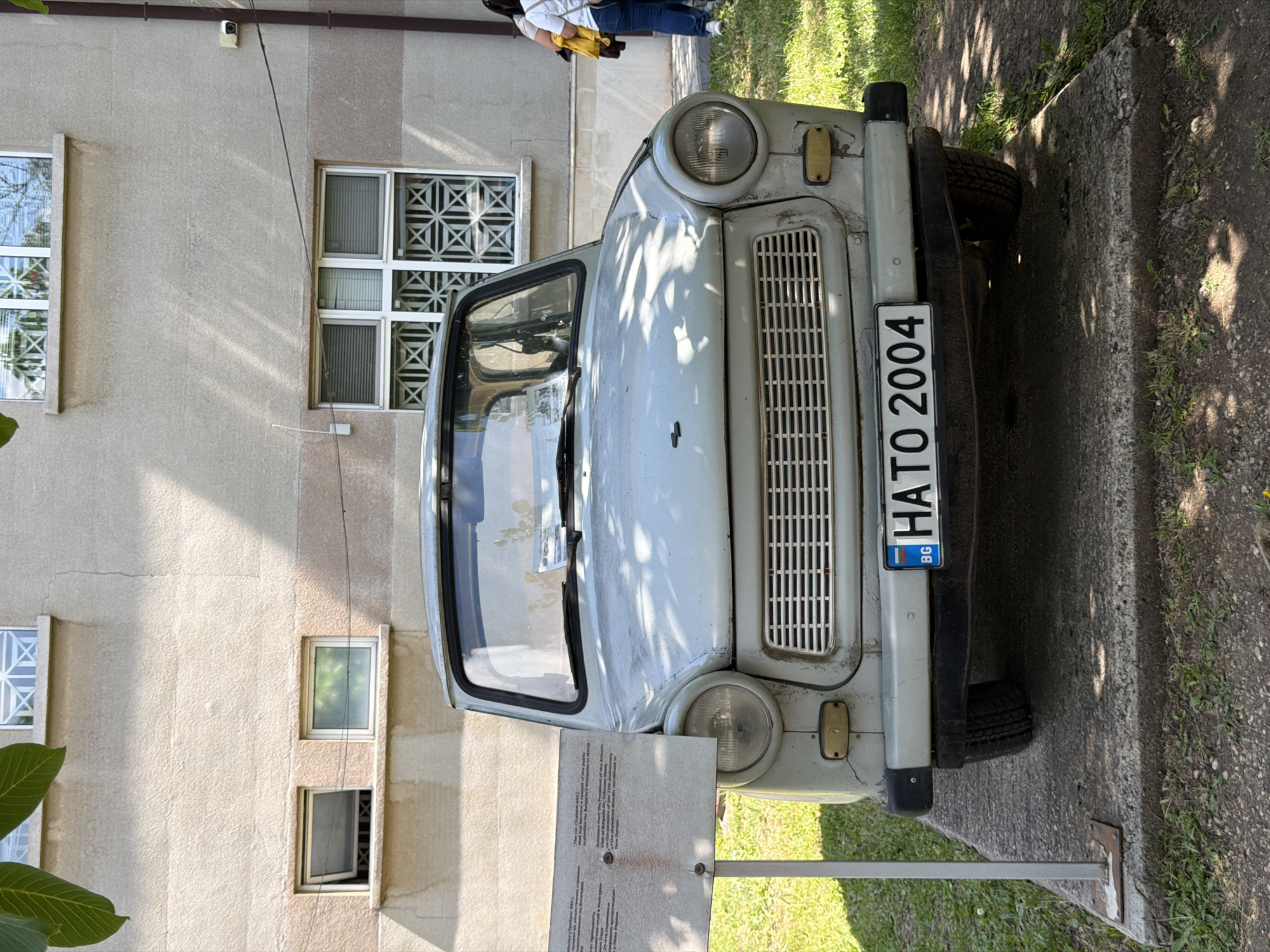 A light blue Trabant 601 is parked on a concrete platform in front of a textured beige building. The car has a license plate that reads "BG HATO 2004". The building has several windows; the upper windows are partially obscured by foliage, and the lower windows are mostly covered with blinds or decorative grilles. A sign is posted to the left of the car. The ground around the concrete platform is covered with grass and dirt. Two women are visible in the background, standing to the right of the car. The scene appears to be outdoors, with shadows from trees falling on the car and ground. The location is the Museum of Socialist Art in Sofia, Bulgaria.